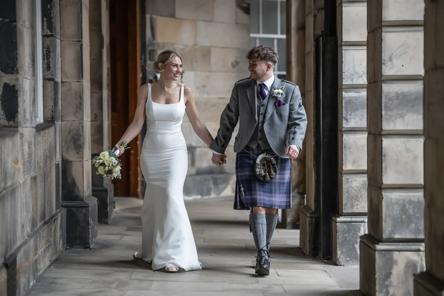 A bride in a white dress and a groom in a kilt walk hand in hand through a stone corridor, both smiling at each other.