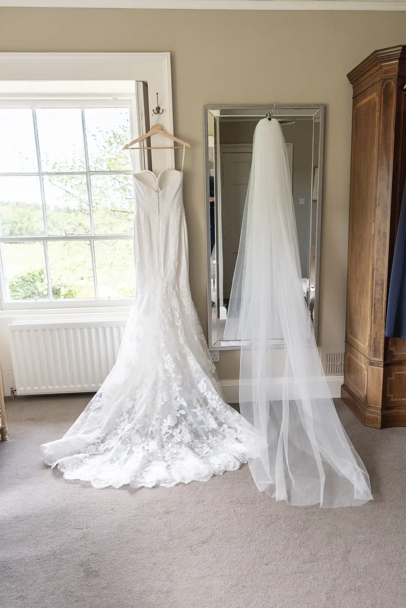 A wedding dress and veil hanging next to a window and a full-length mirror in a bright room.