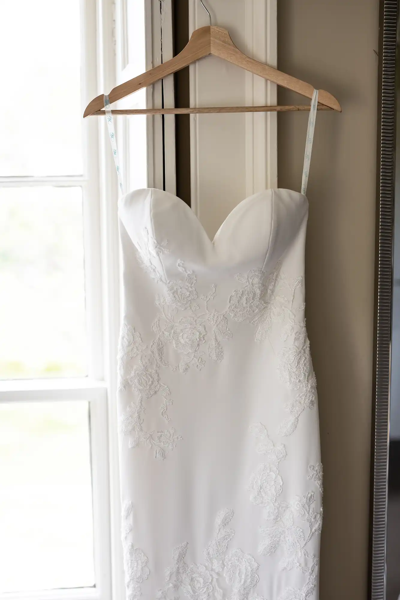 A white wedding dress with lace details hanging on a wooden hanger in front of a window.