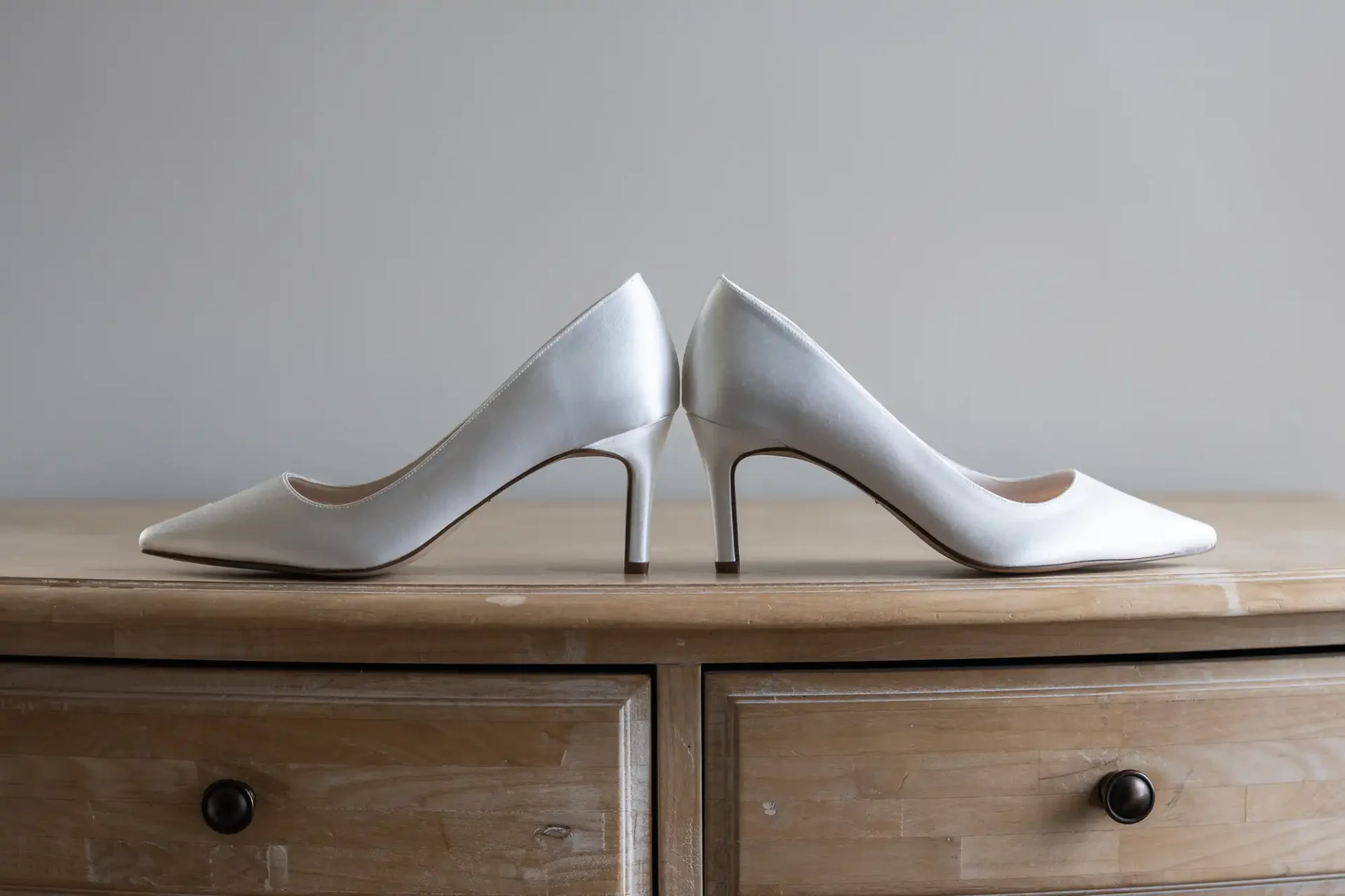 A pair of elegant white high-heeled shoes positioned back-to-back on a wooden dresser against a neutral background.
