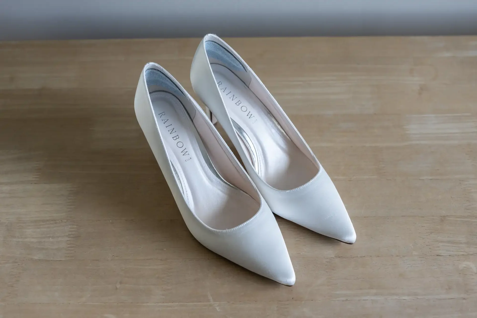A pair of white satin bridal shoes on a wooden floor.