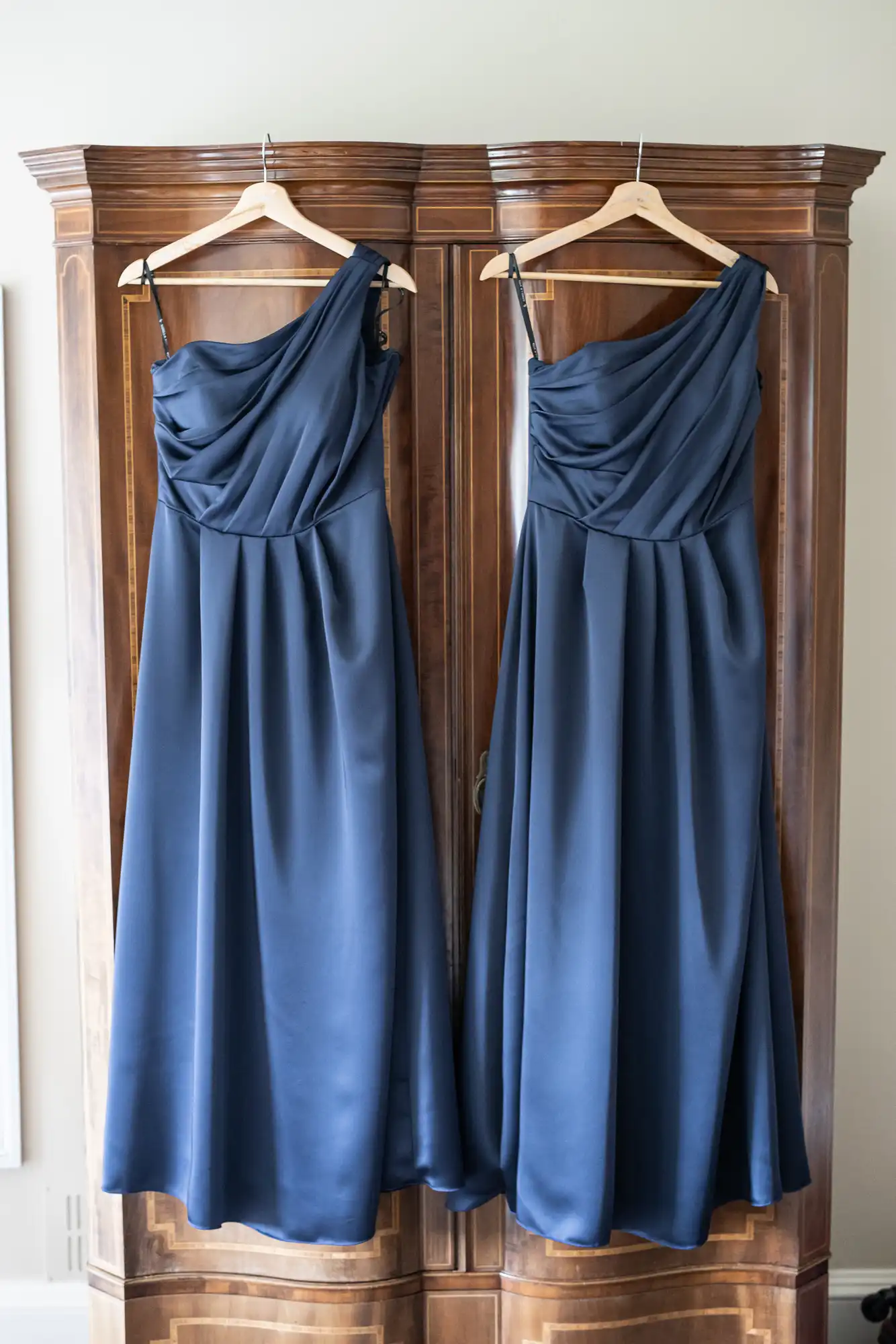 Two blue bridesmaid dresses hanging on wooden hangers against an antique wooden wardrobe.