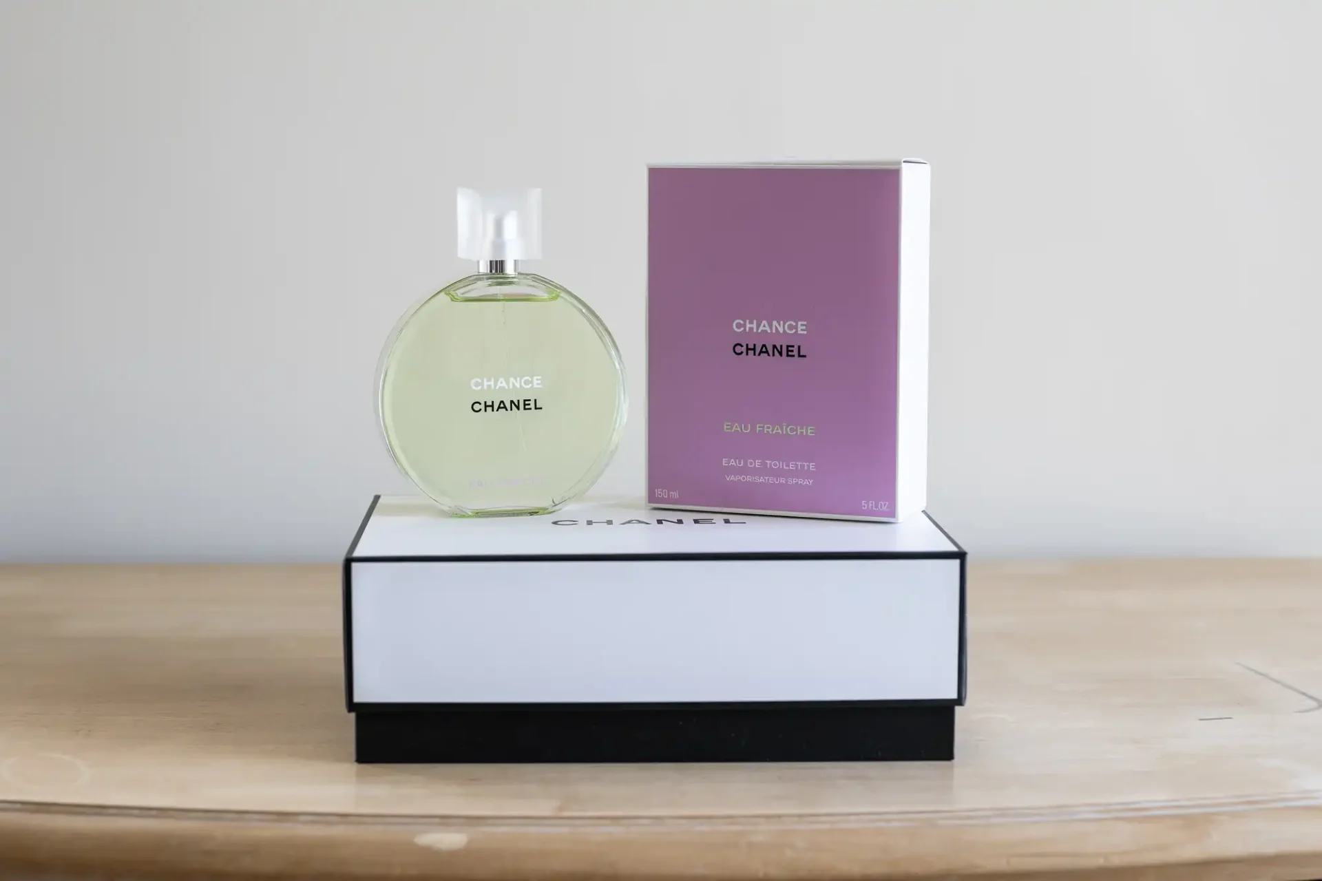 A round bottle of chanel chance perfume next to its purple and white packaging box, placed on a black platform with a wooden surface background.