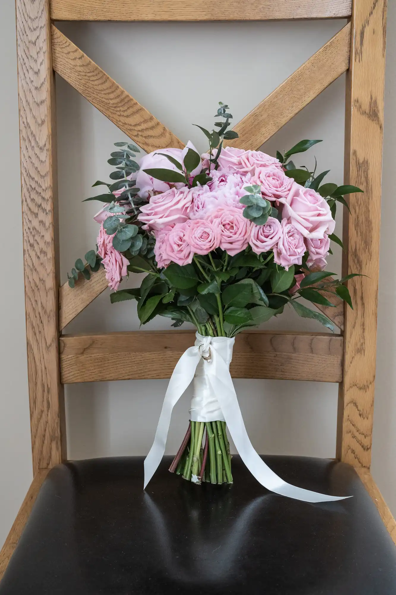 A bouquet of pink roses tied with a white ribbon, resting on a wooden chair against a beige wall.