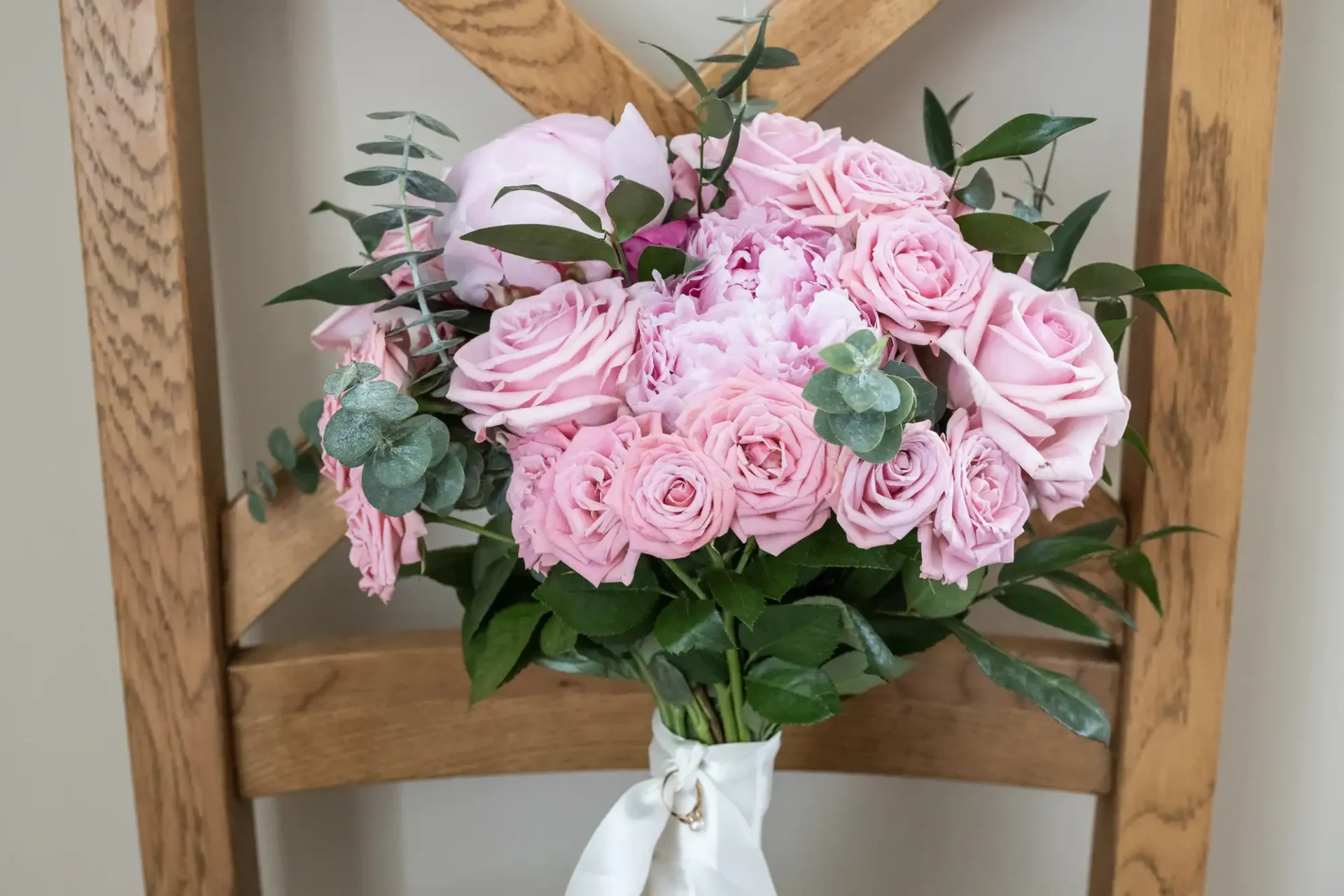 A bouquet of pink roses and greenery tied with a white ribbon, resting on a wooden chair.