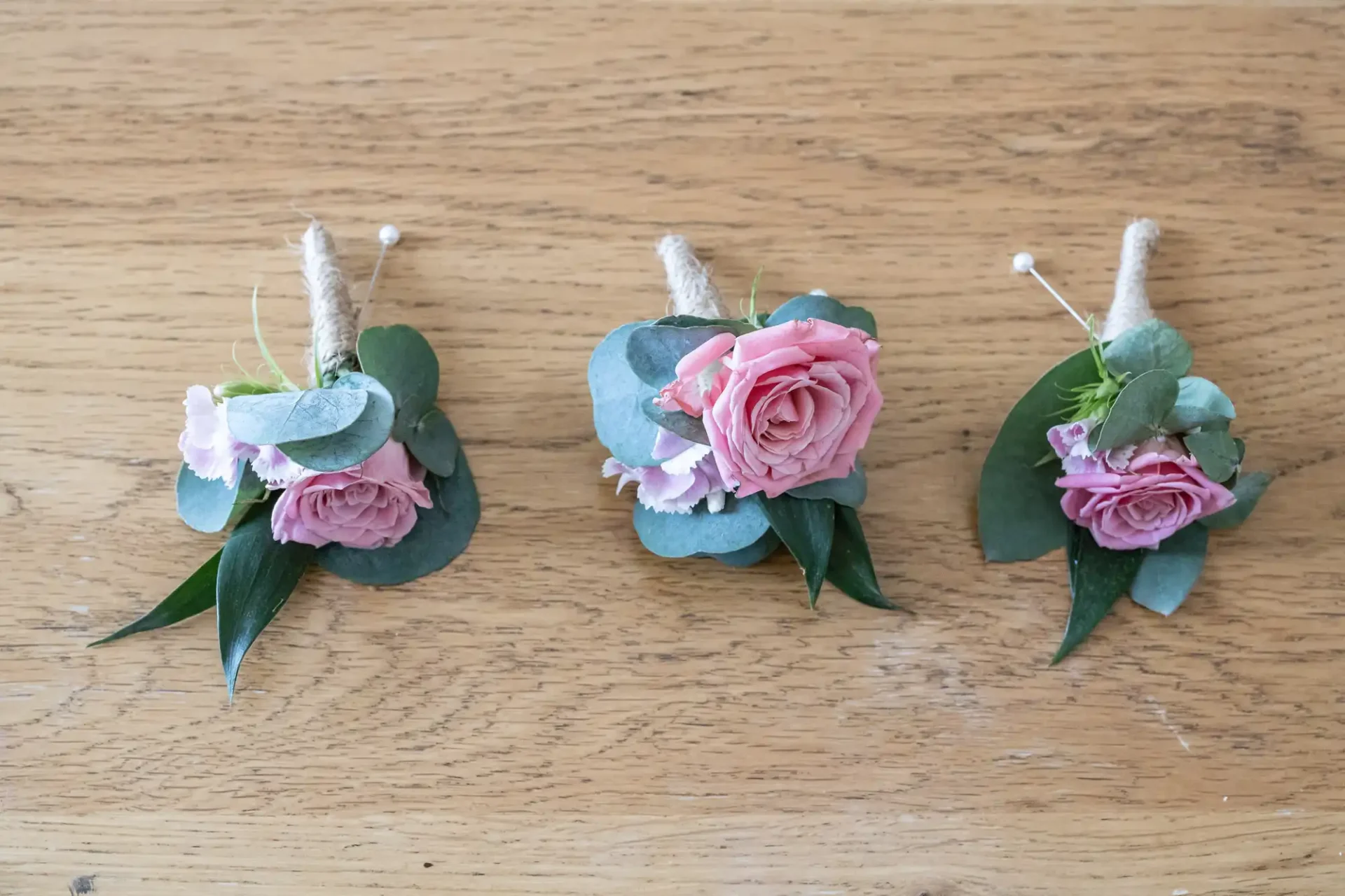 Three pink rose boutonnieres with eucalyptus leaves and twine on a wooden surface.