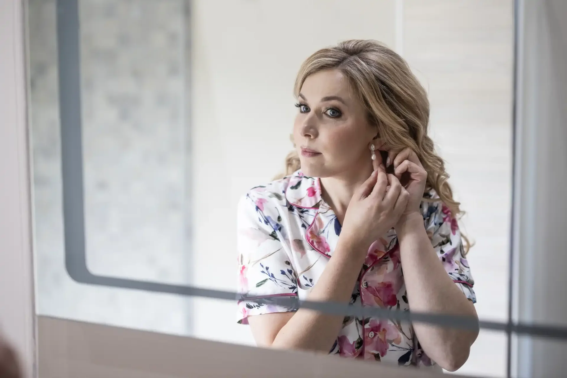 A woman with blonde hair wearing a floral blouse gazes thoughtfully through a window while adjusting her earring.