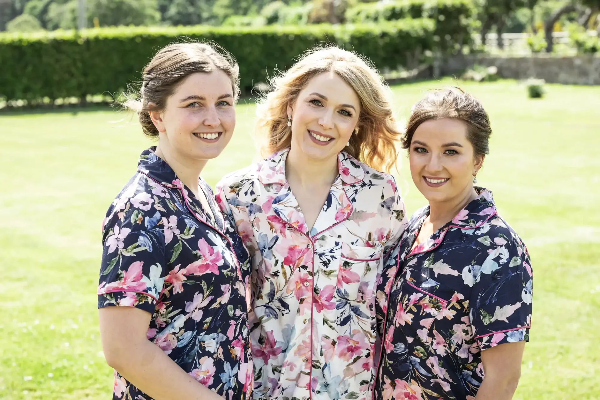 Three women in floral dresses smiling outdoors on a sunny day.