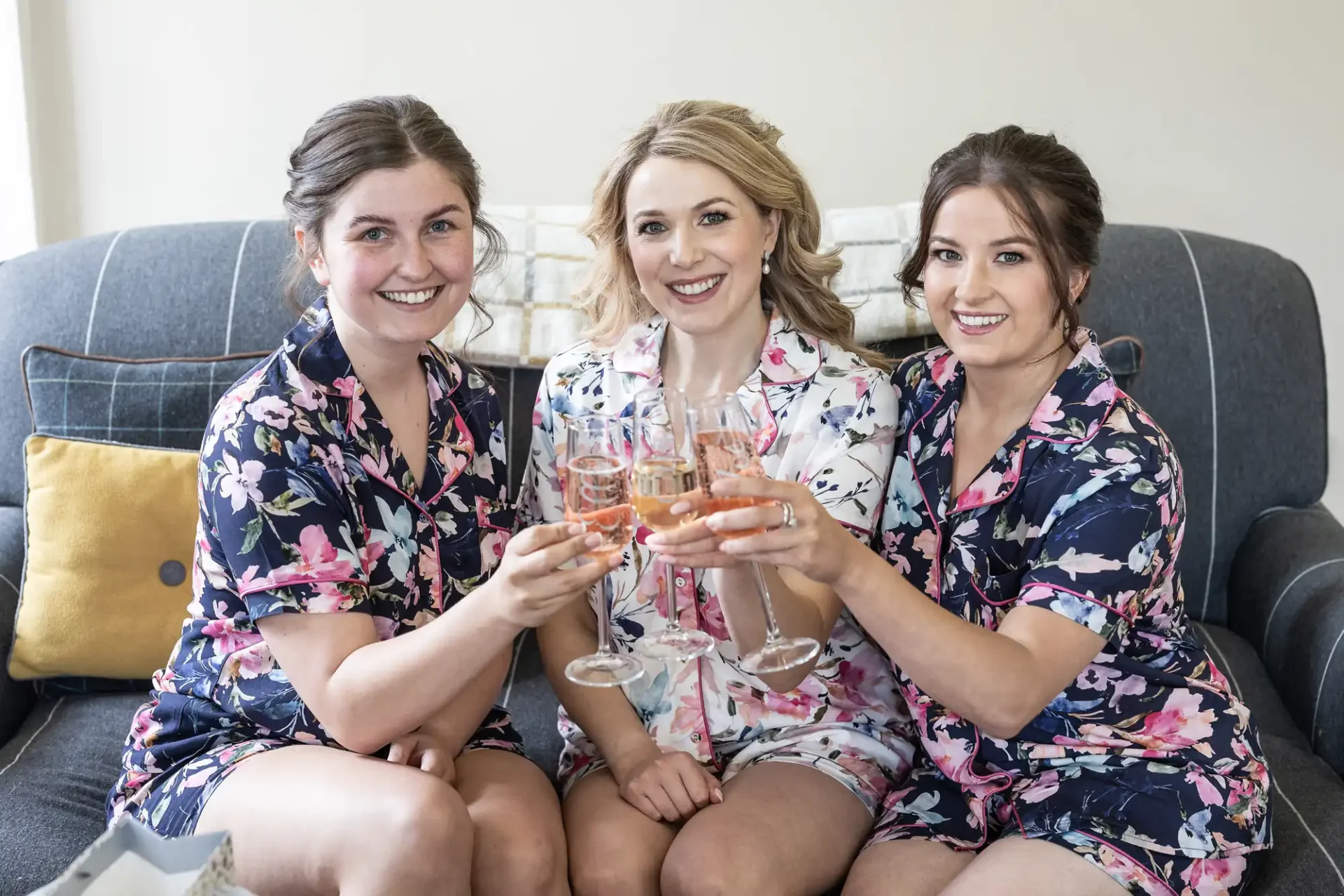 Three women in floral robes sitting on a couch, smiling and toasting with glasses of rosé wine.
