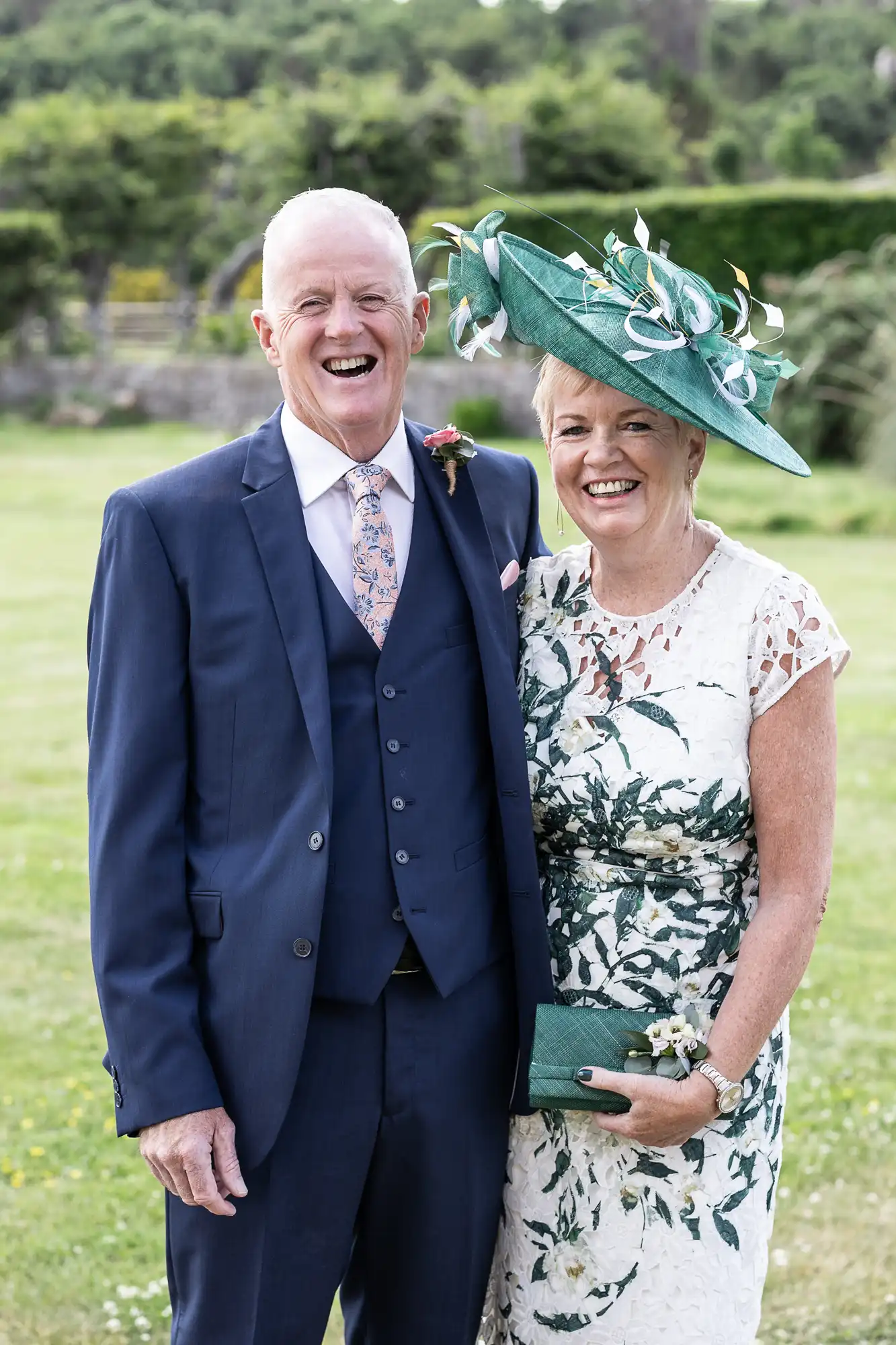 An older couple in formal wear smiling at a wedding event; the man in a navy suit and the woman in a floral dress and a large green hat.