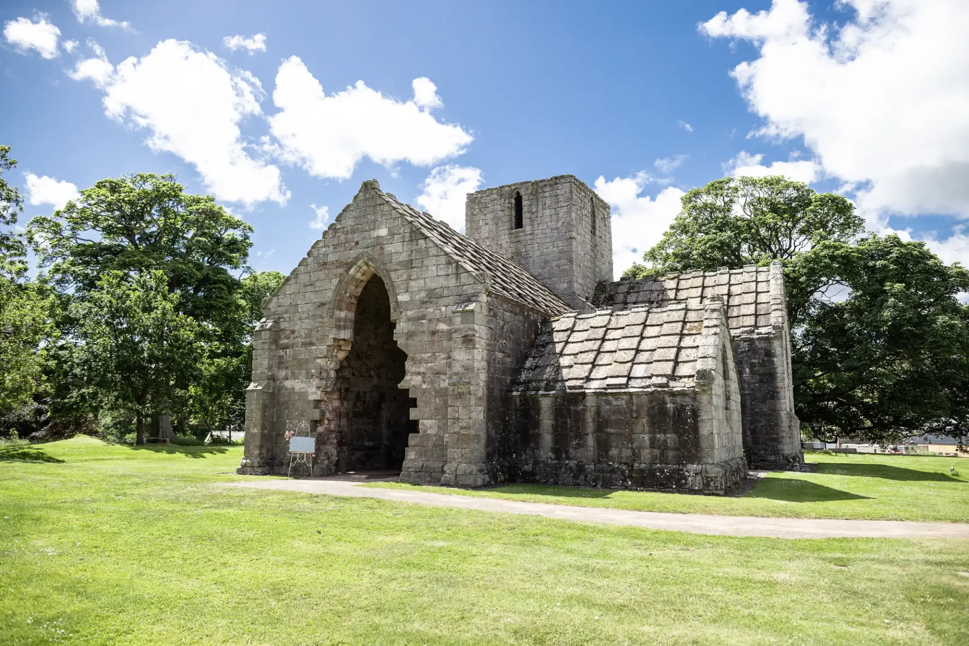 Stone ruins of a medieval abbey with an arched entrance, surrounded by lush green grass and trees under a clear blue sky.