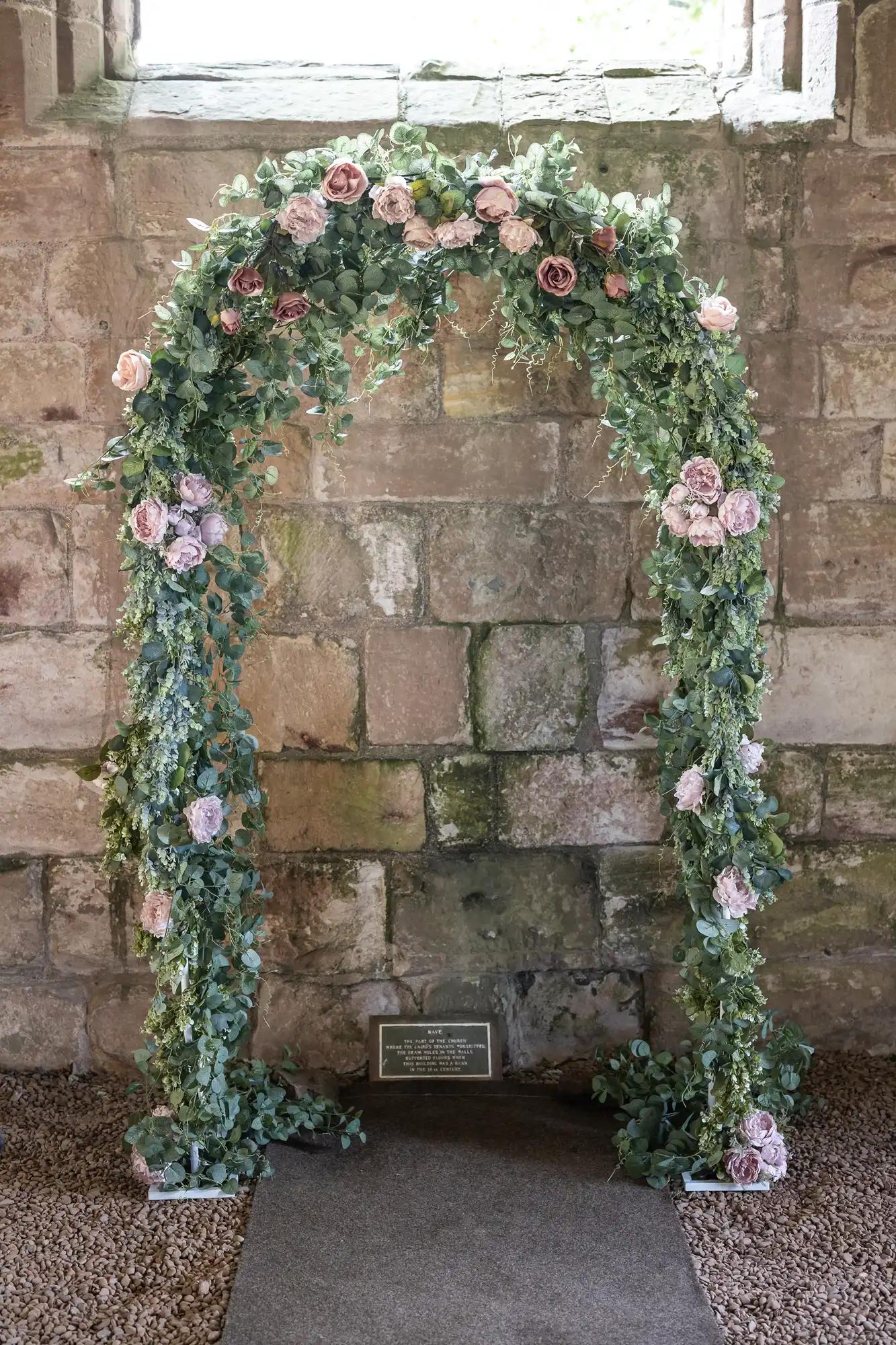 Floral arch with pink and white roses and green leaves, set against an aged stone wall in a historic building.