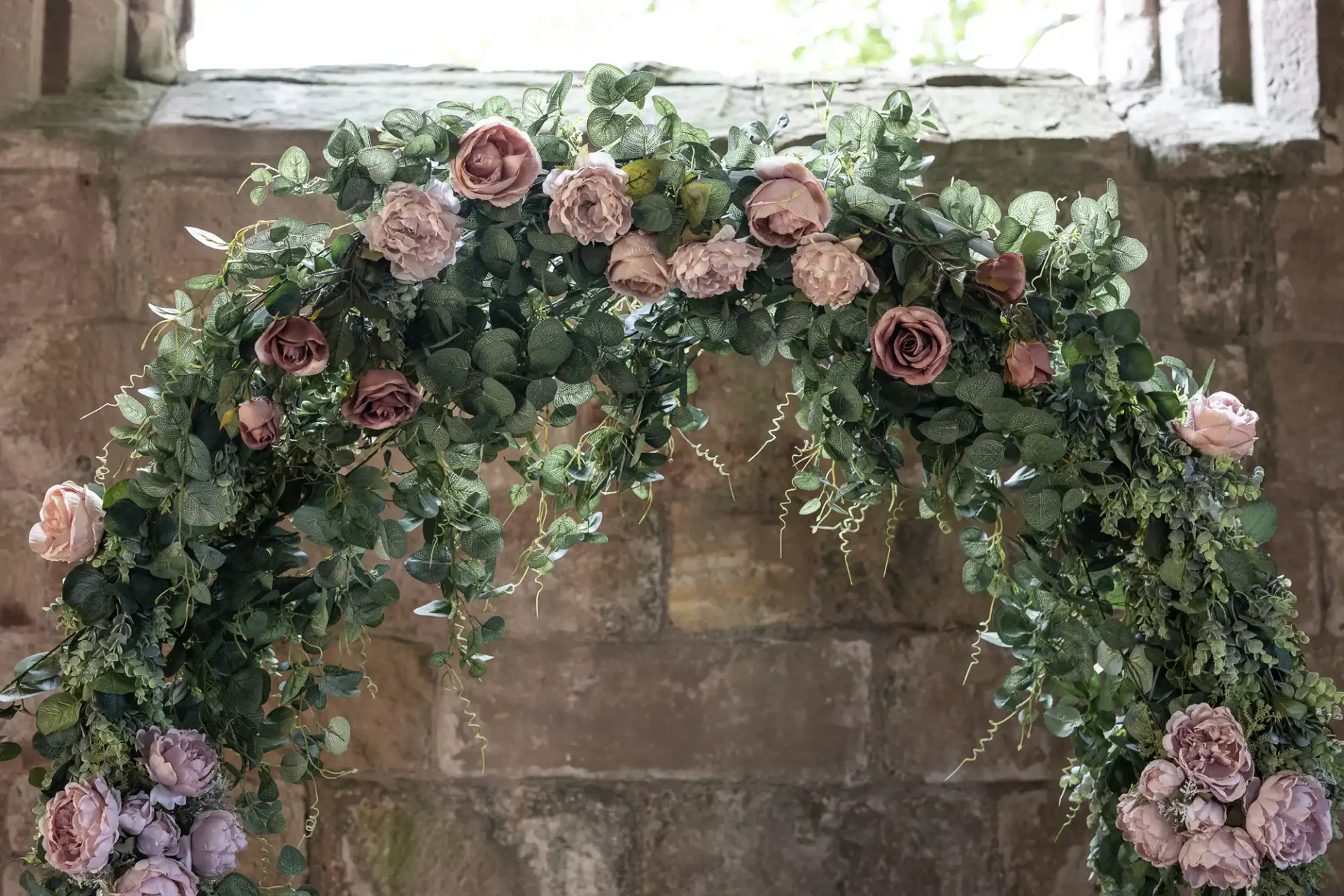 Arch-shaped floral arrangement with pink and cream roses integrated with green ivy leaves, set against a stone wall background.