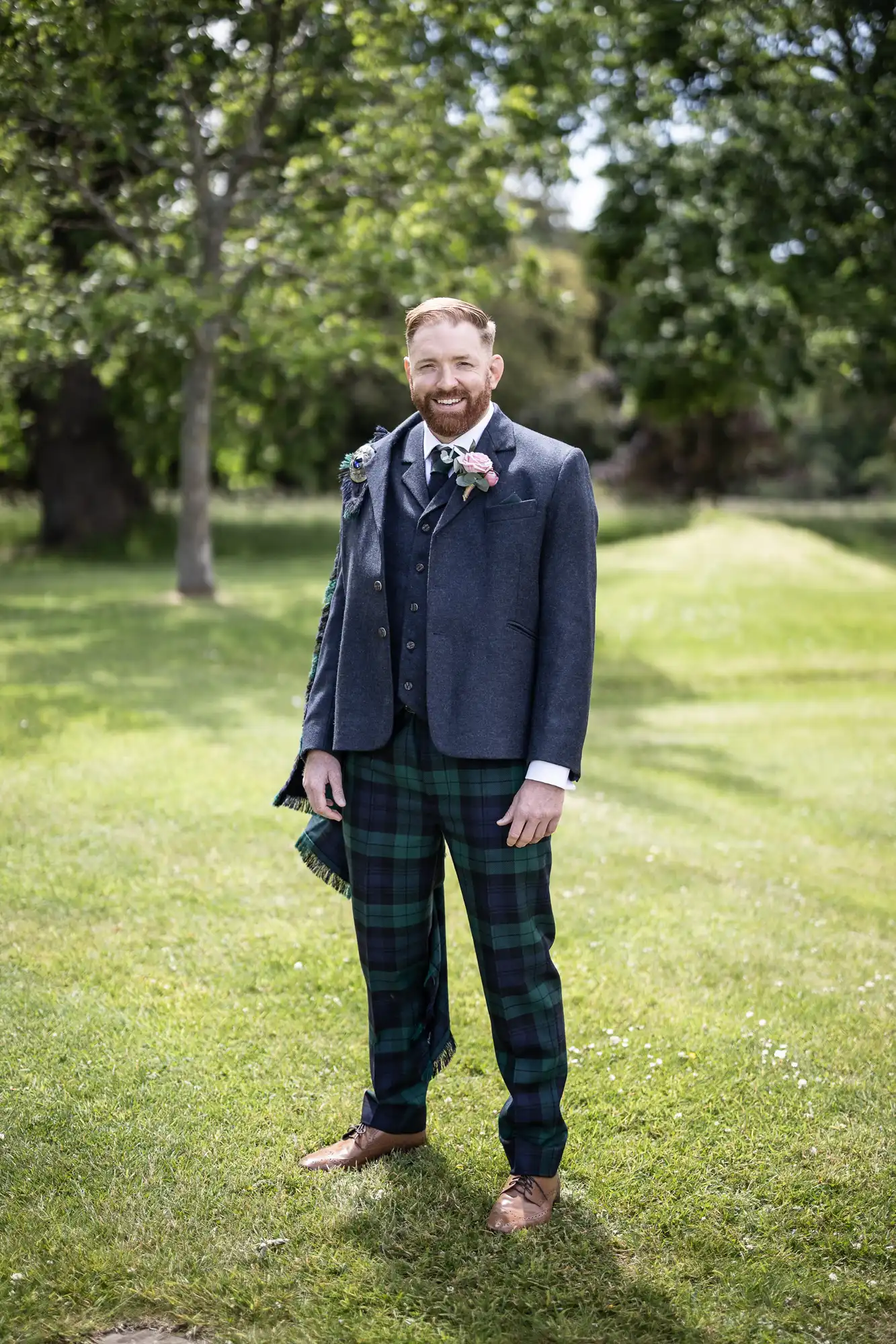 A man in a blue blazer and green tartan trousers stands smiling in a sunny park, holding a hat and wearing a boutonniere.