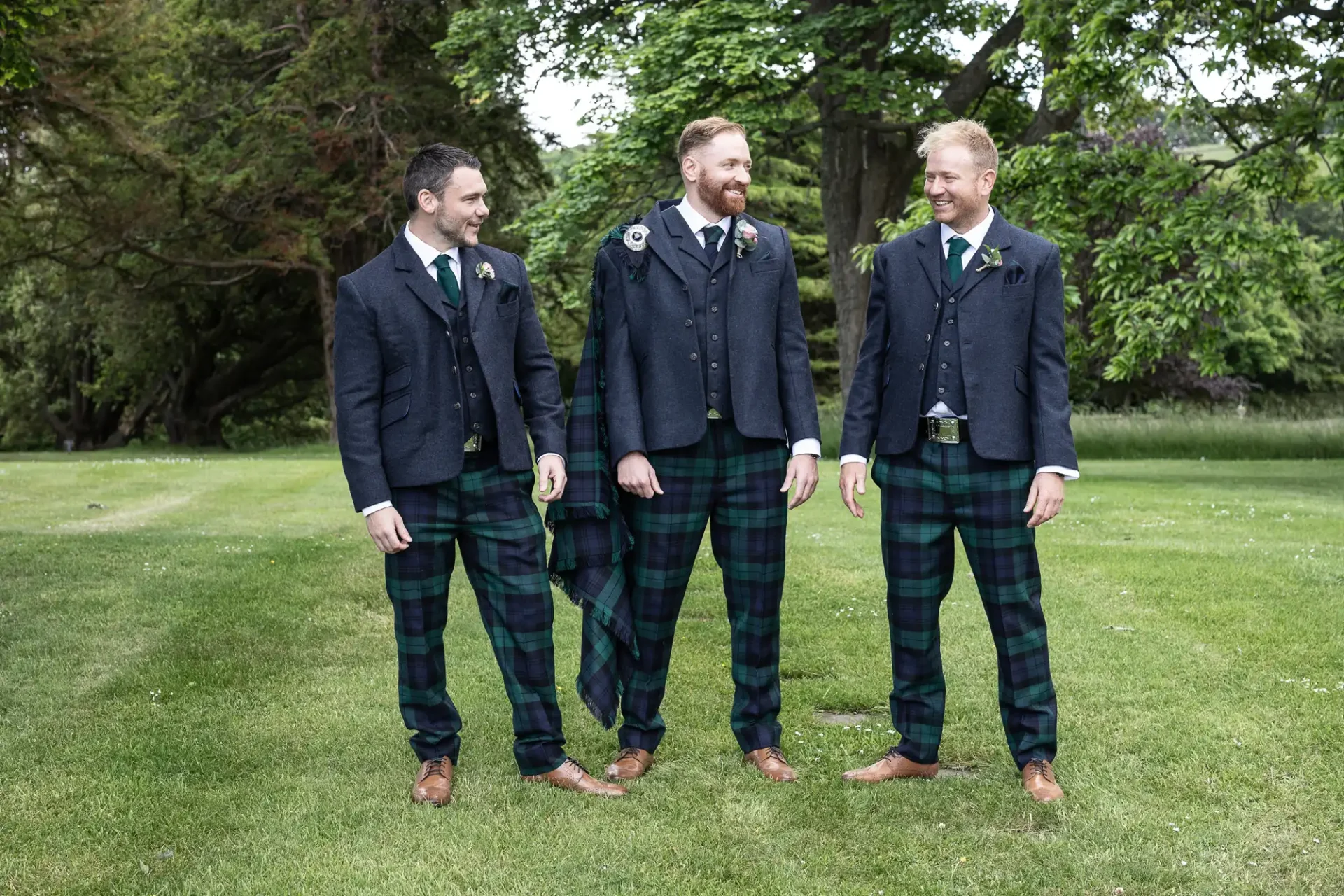 Three men in traditional tartan kilts and dark jackets smiling and standing in a lush green garden.