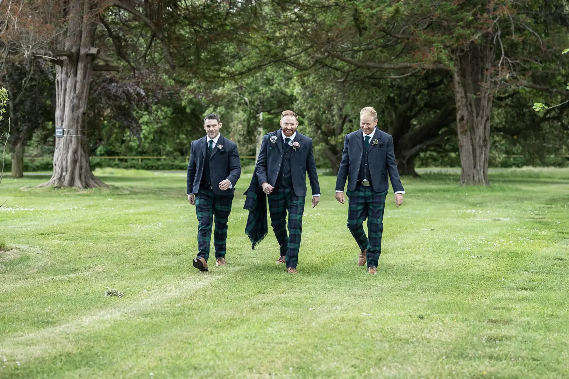 Three men in suits and tartan kilts walking joyfully through a grassy park with trees in the background.