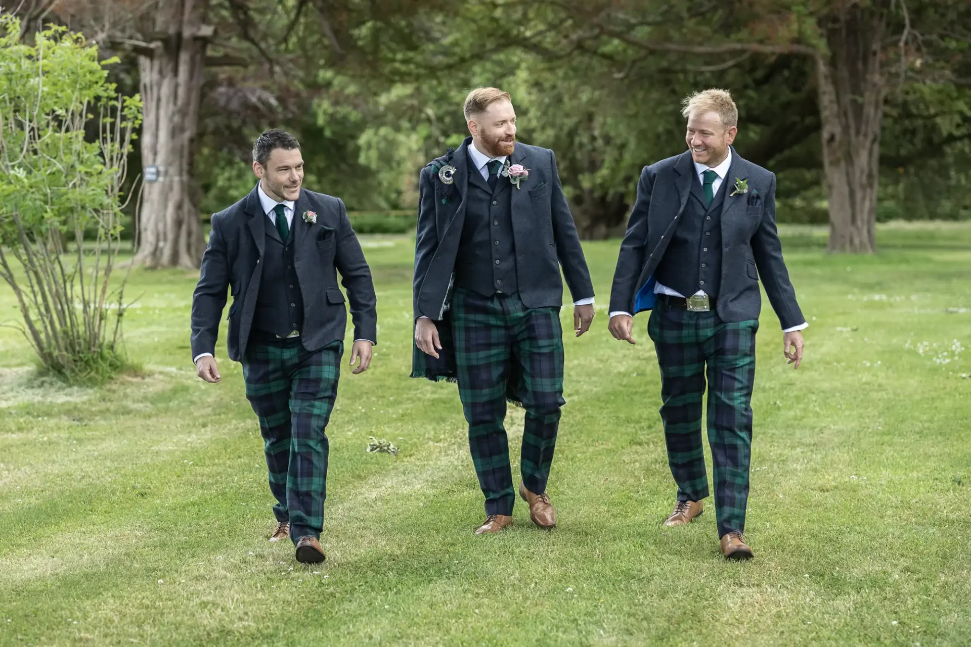 Three men in matching tartan kilts and jackets walk across a grassy area, laughing and enjoying each other's company.