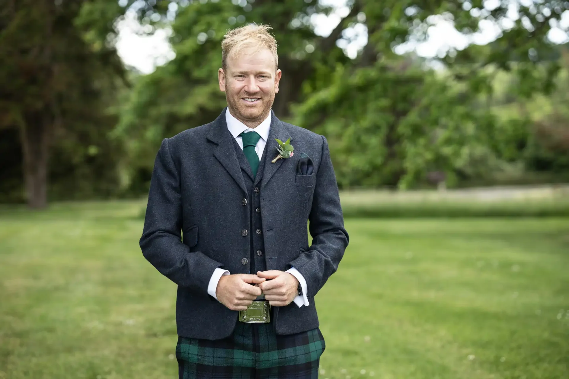 A man in a traditional scottish kilt and tweed jacket smiles outdoors, holding a glass, with a boutonniere on his lapel.