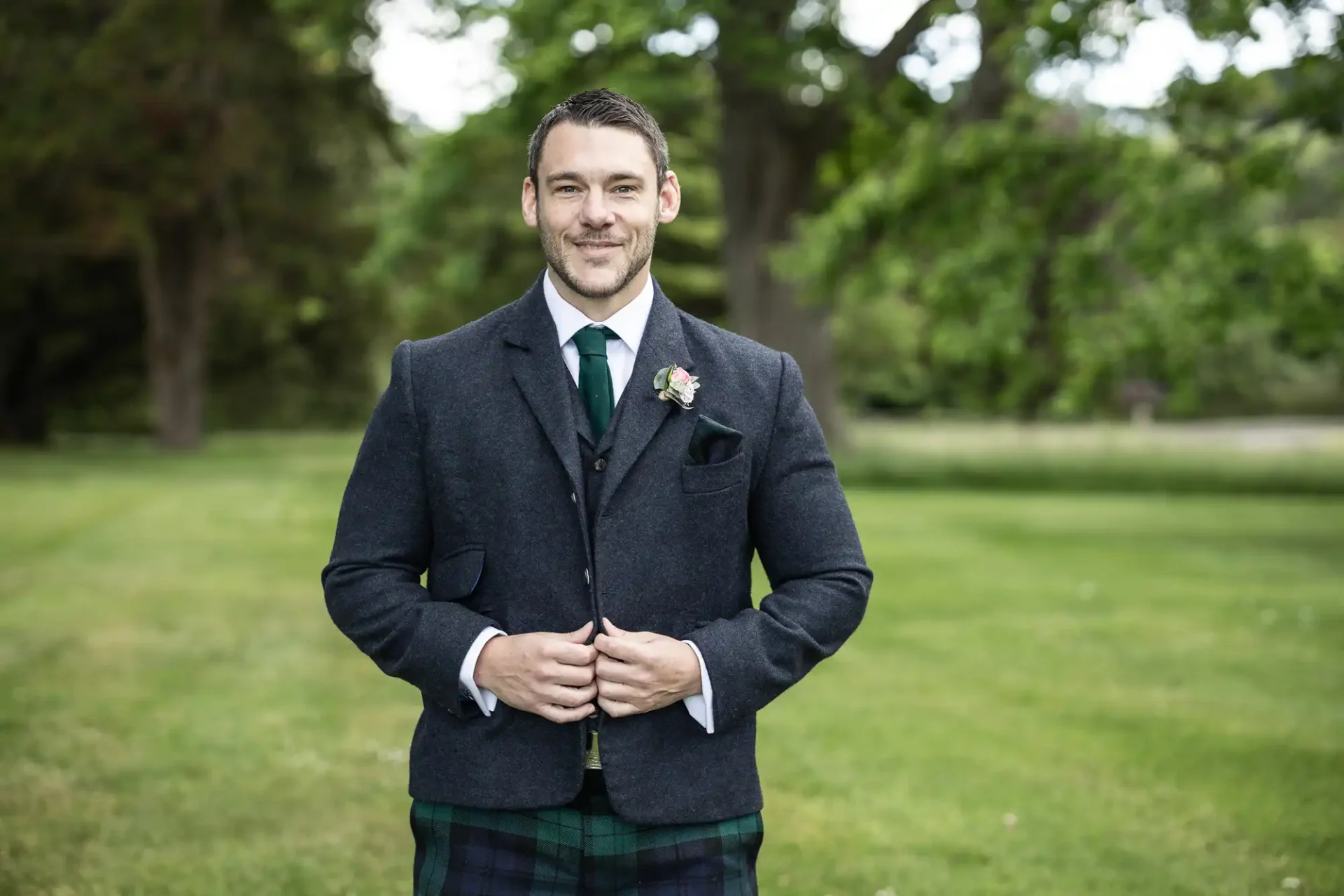 Man in traditional scottish attire, including a kilt and tweed jacket, smiling in a lush garden setting.