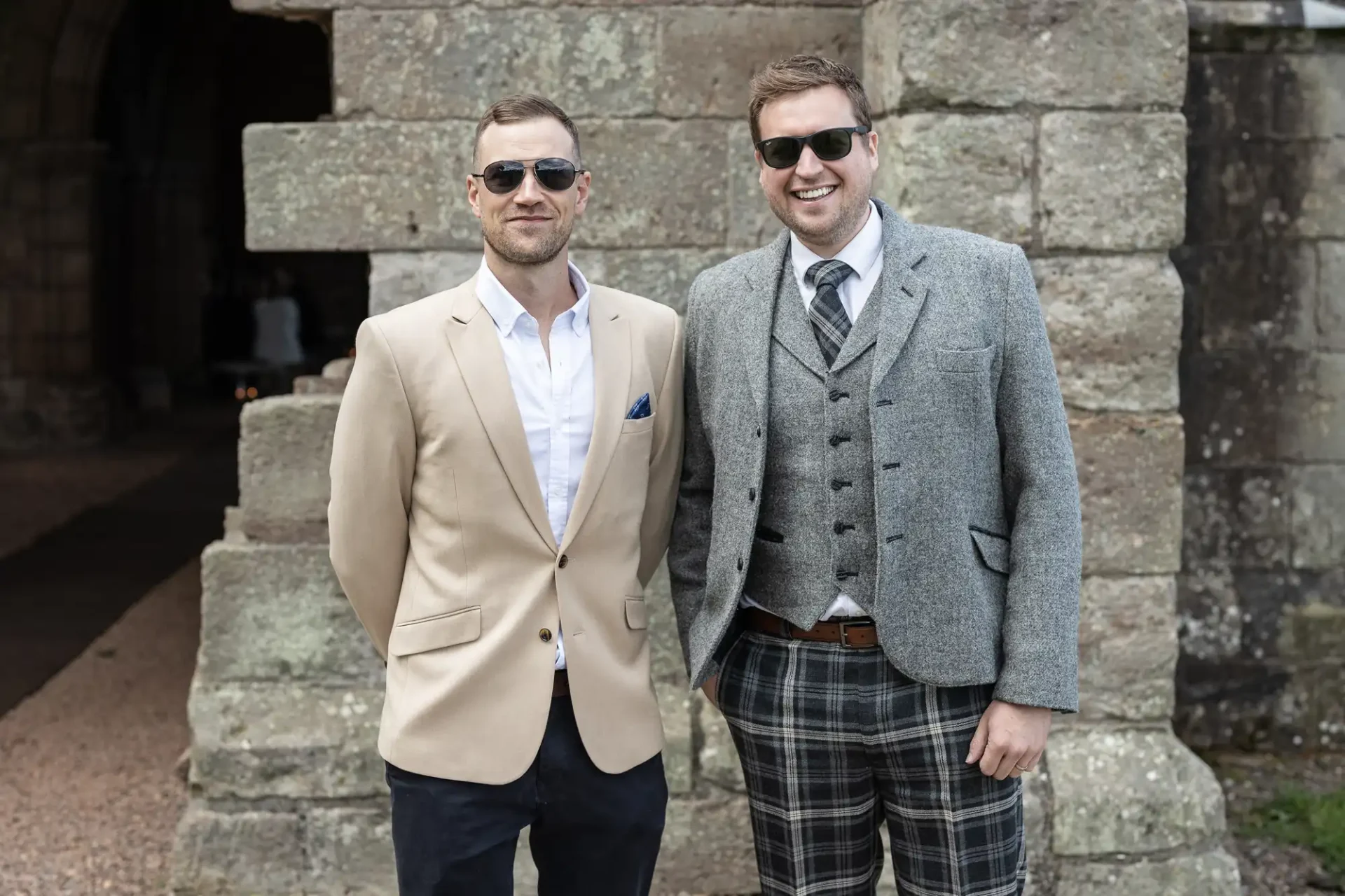 Two men smiling, one in a tan blazer and sunglasses, the other in a gray tweed jacket and plaid trousers, standing in front of a stone wall.