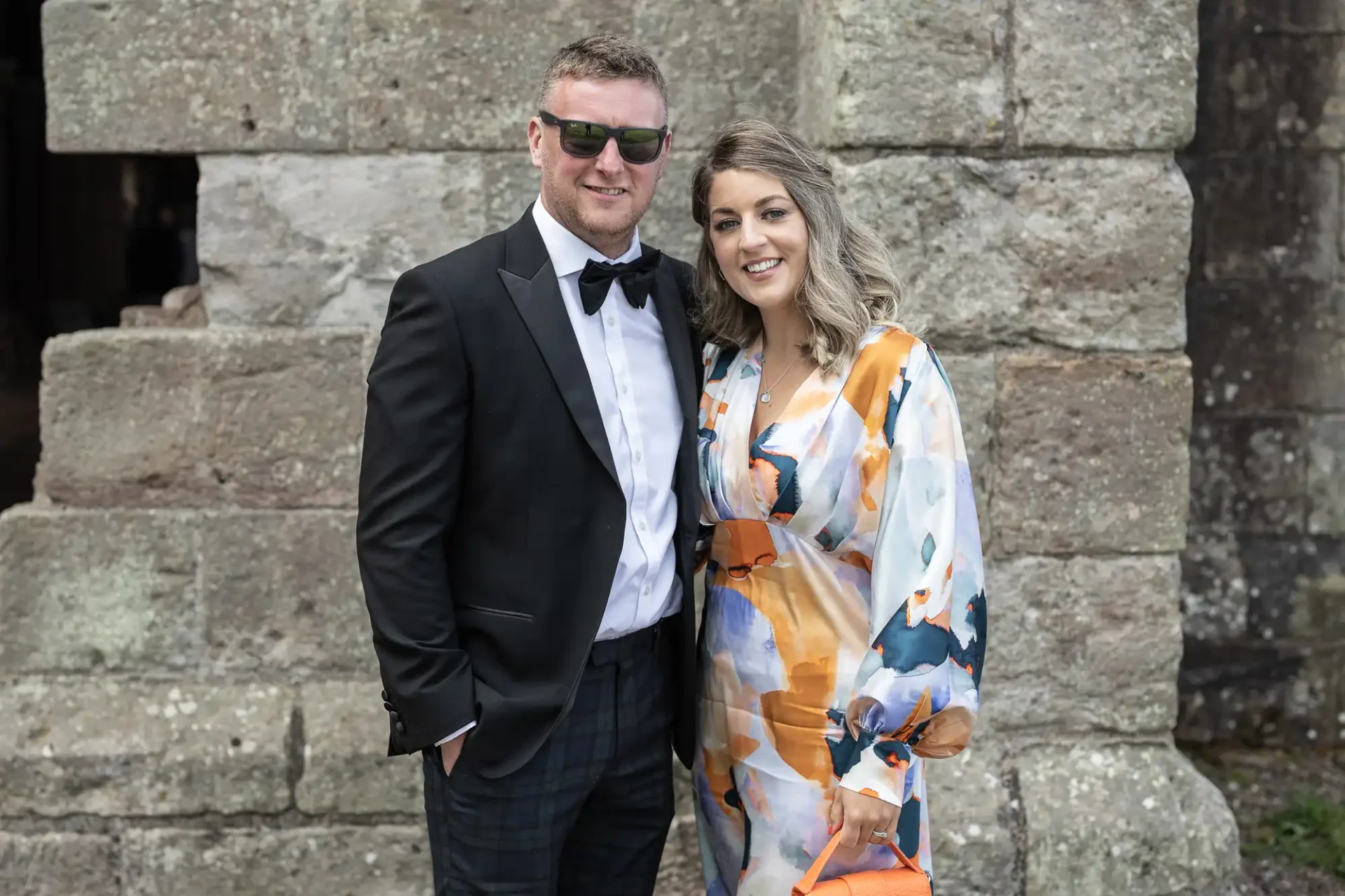 A man in a black tuxedo and sunglasses stands next to a woman in a colorful floral dress, both smiling in front of a stone structure.
