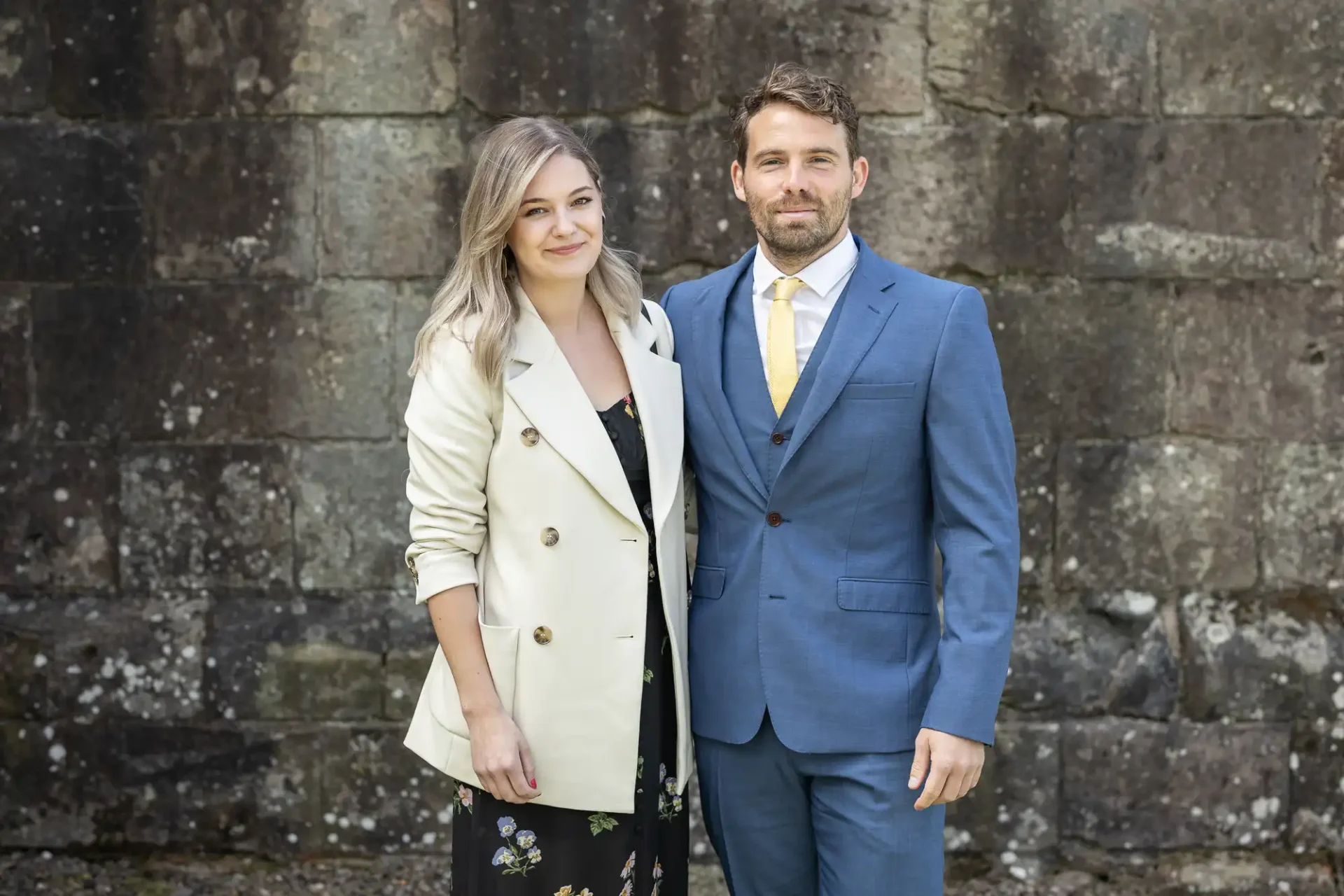 A man in a blue suit and yellow tie and a woman in a cream blazer and floral dress standing together in front of a stone wall.
