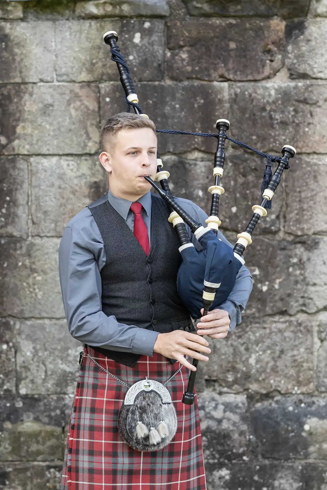 A young man in traditional scottish attire, including a red tartan kilt and sporran, playing the bagpipes.
