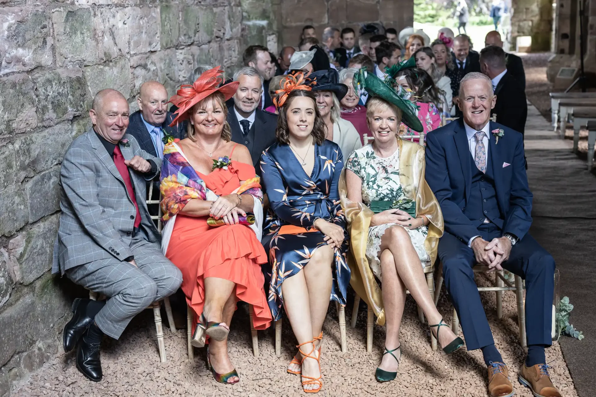 A group of elegantly dressed guests, some wearing elaborate hats, seated together at a wedding ceremony in a stone-walled venue.