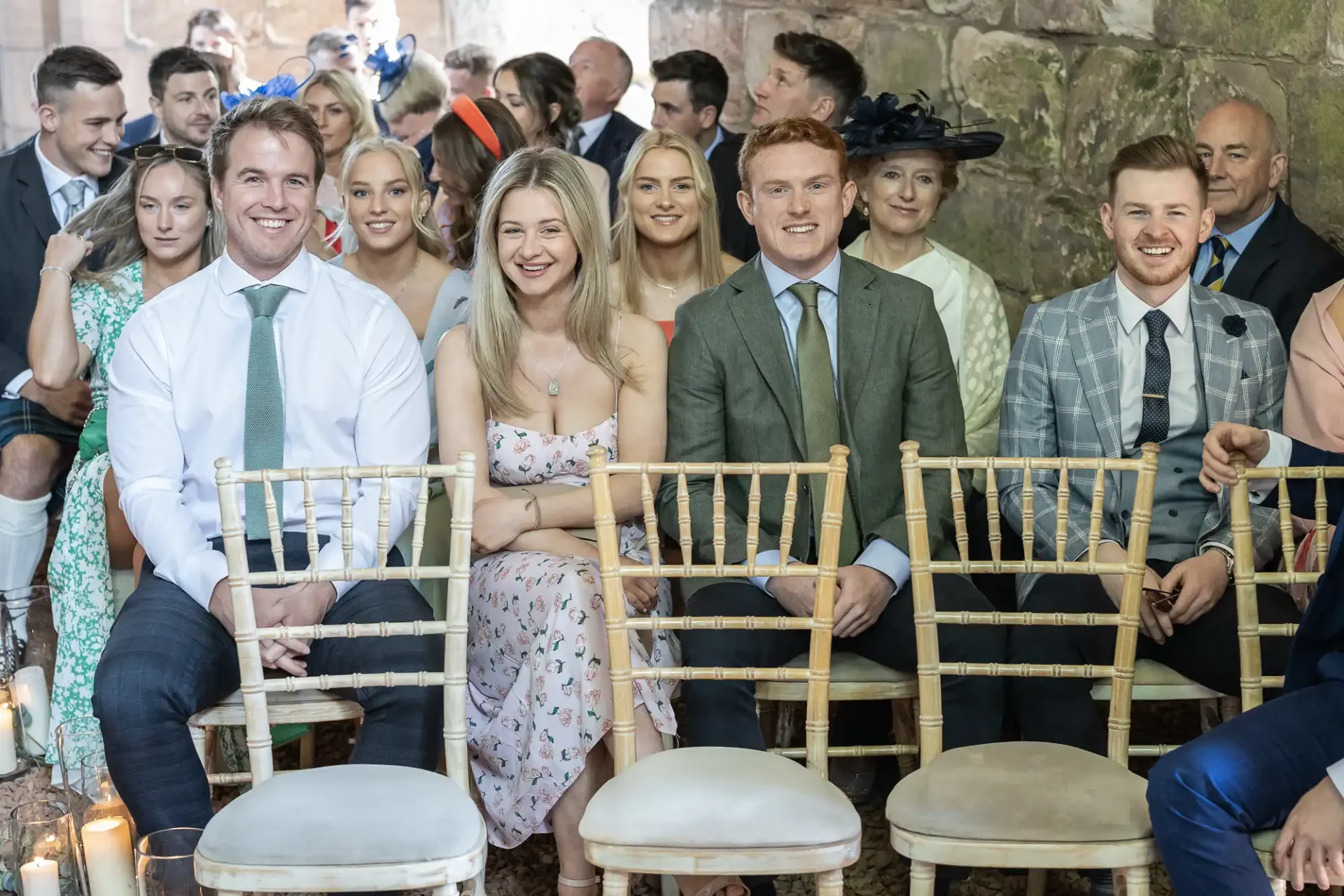 Guests smiling and sitting in rows at a wedding ceremony inside a stone-walled venue with wooden chairs and candle decorations.