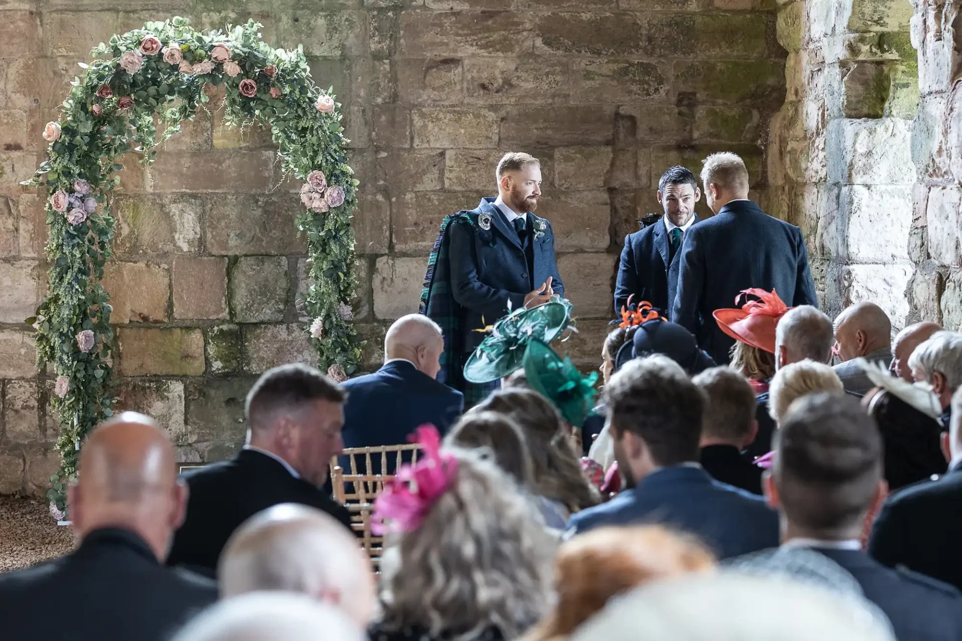A wedding ceremony in a rustic building with stone walls, featuring guests in formal attire and a floral arch at the front.