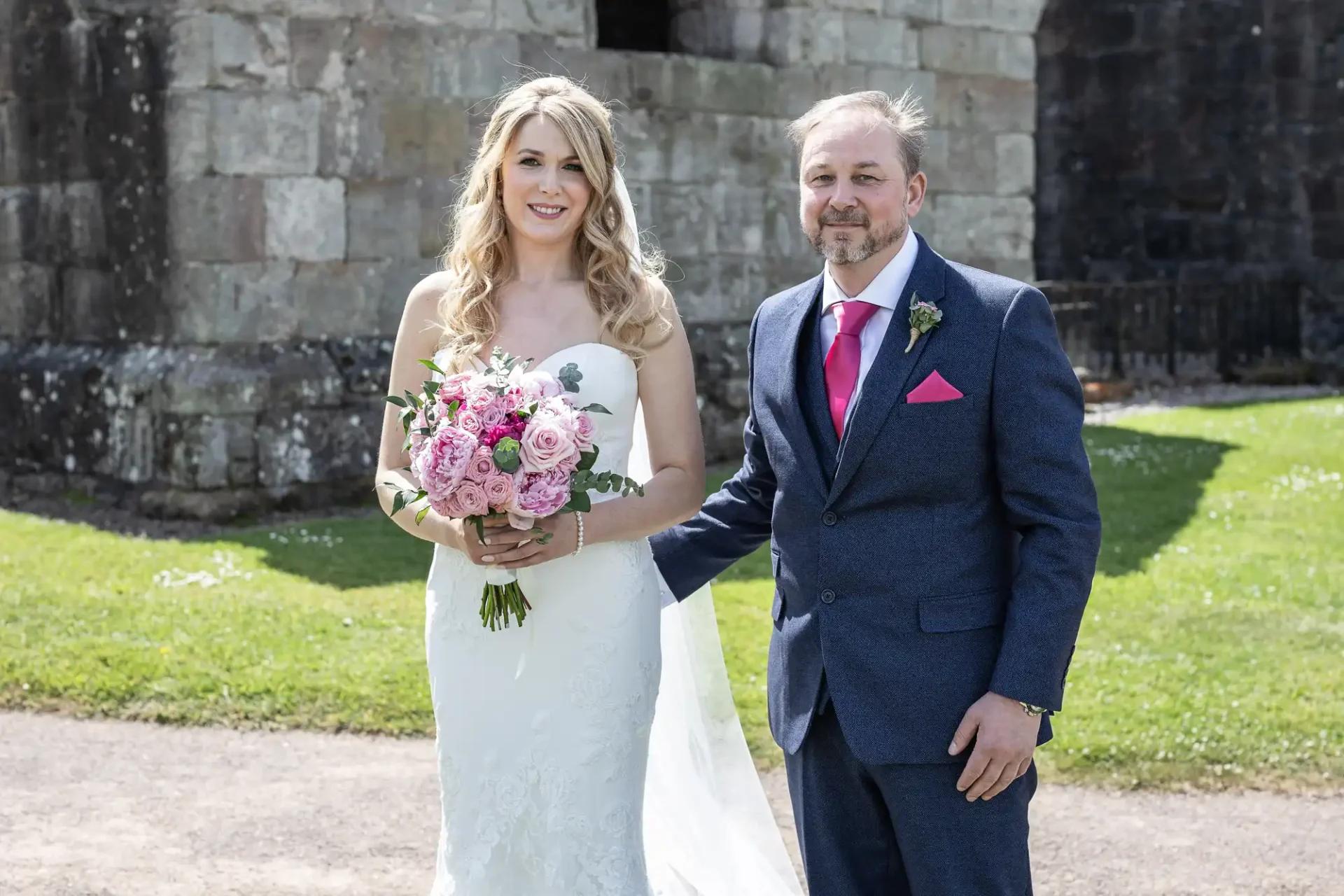 A bride and groom smiling in formal wedding attire, holding hands in front of a stone building, with the bride holding a bouquet of pink flowers.
