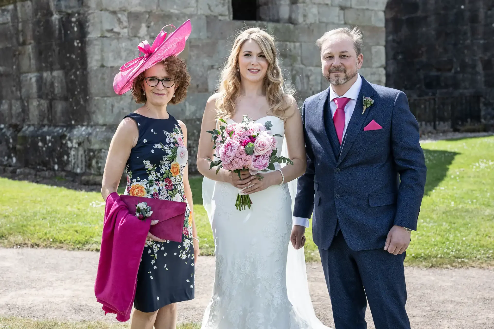 A bride and groom posing with an older woman in a pink hat at a sunny outdoor wedding ceremony.