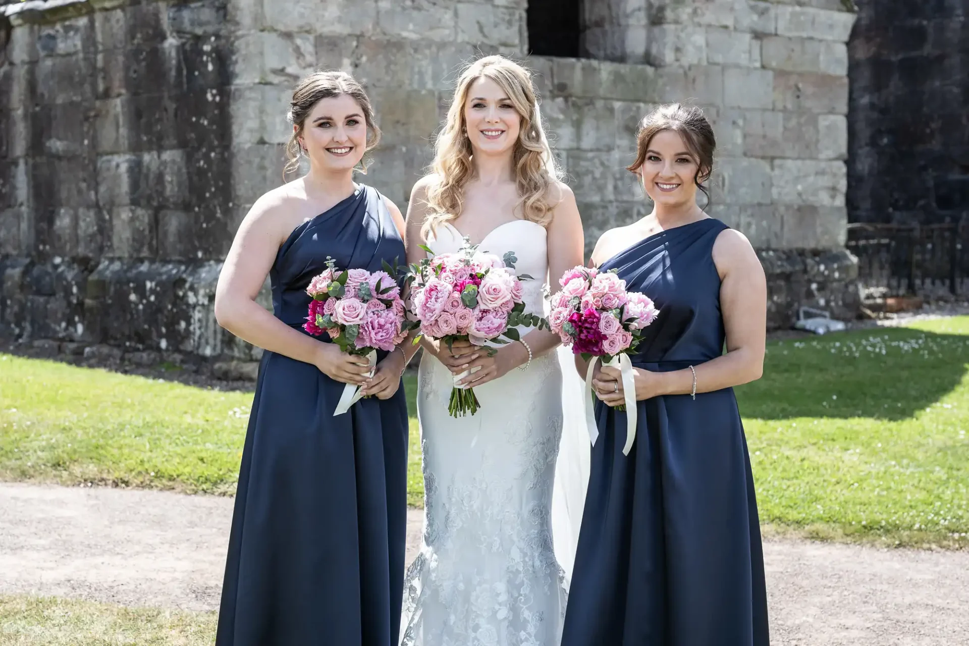 A bride in a white dress stands with two bridesmaids in navy blue dresses, all holding bouquets, in front of a stone building.