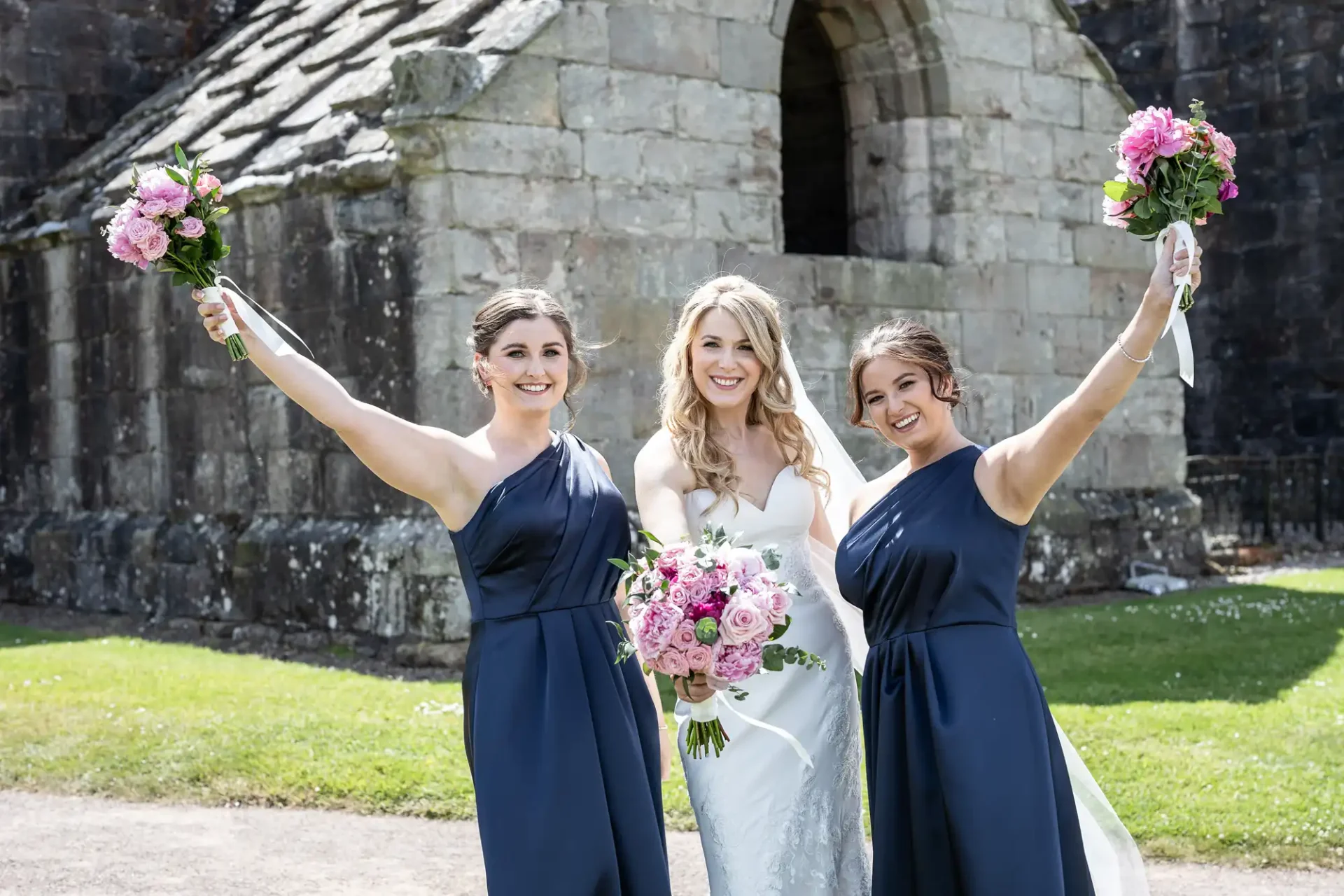 A bride in a white dress and two bridesmaids in navy blue dresses holding pink bouquets, standing before an ancient stone church on a sunny day.