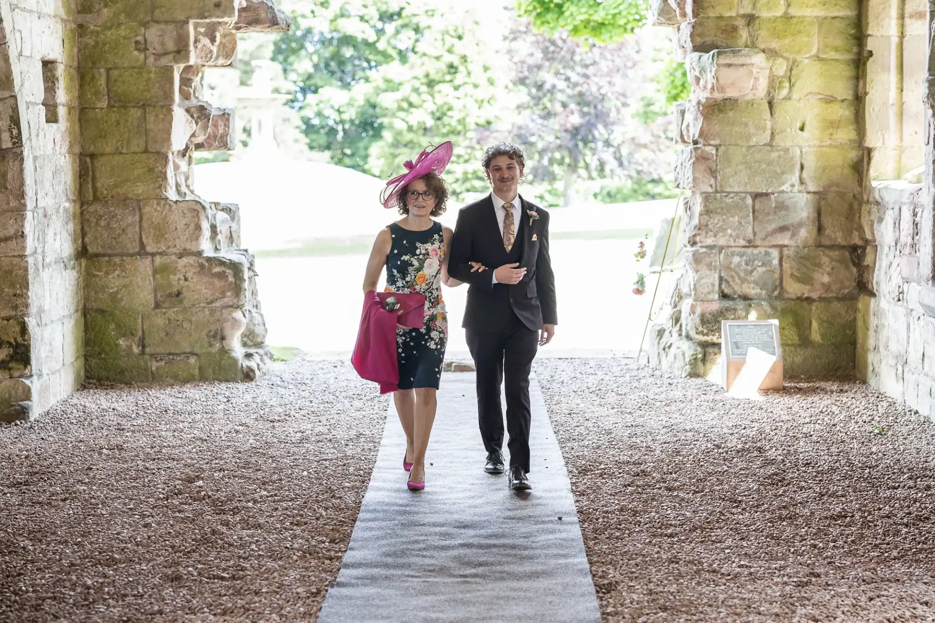 A woman in a pink hat and floral dress and a man in a black suit walk together on a gravel path under an archway.