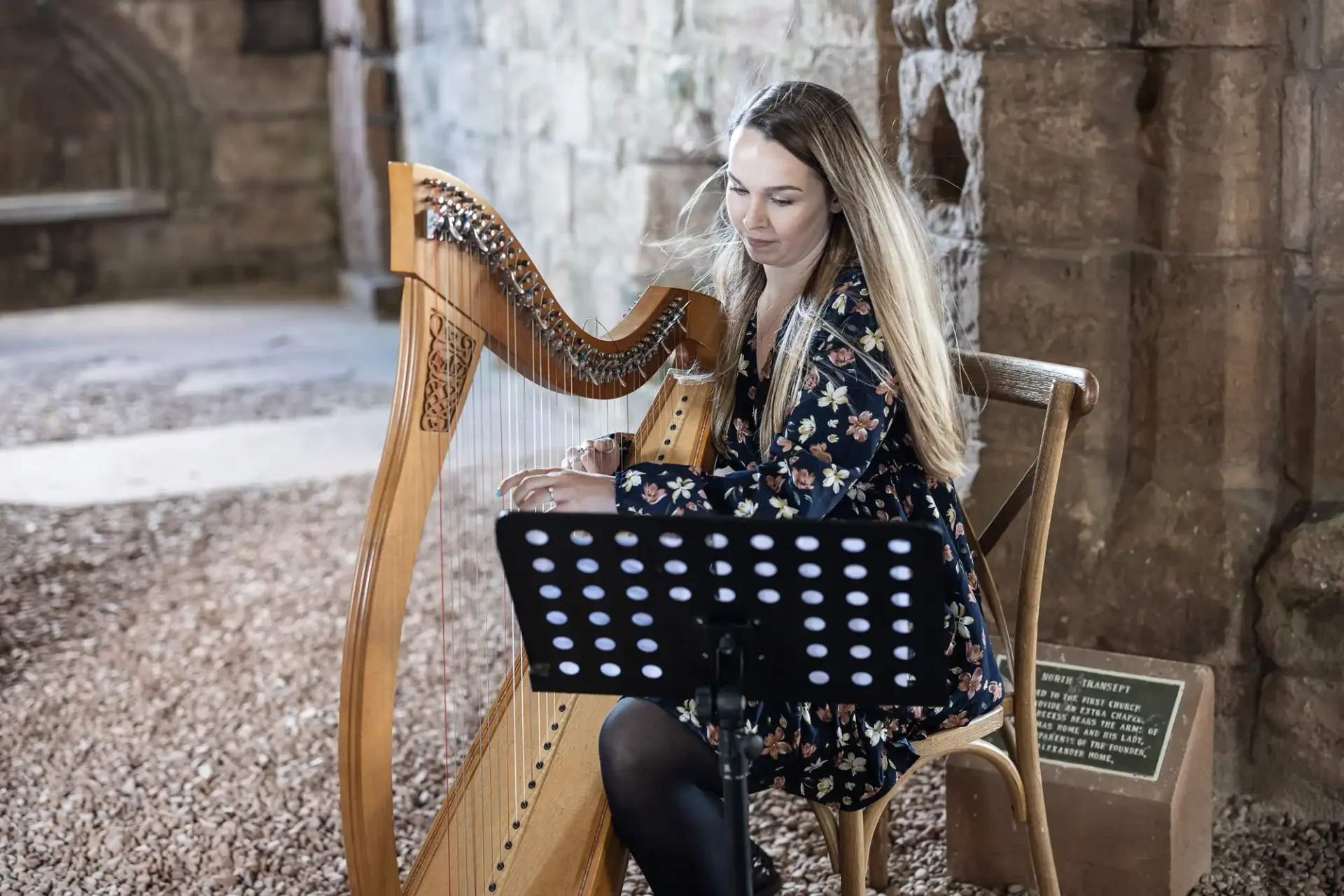 A woman plays a harp inside a historic stone building, focused on sheet music on a black stand.