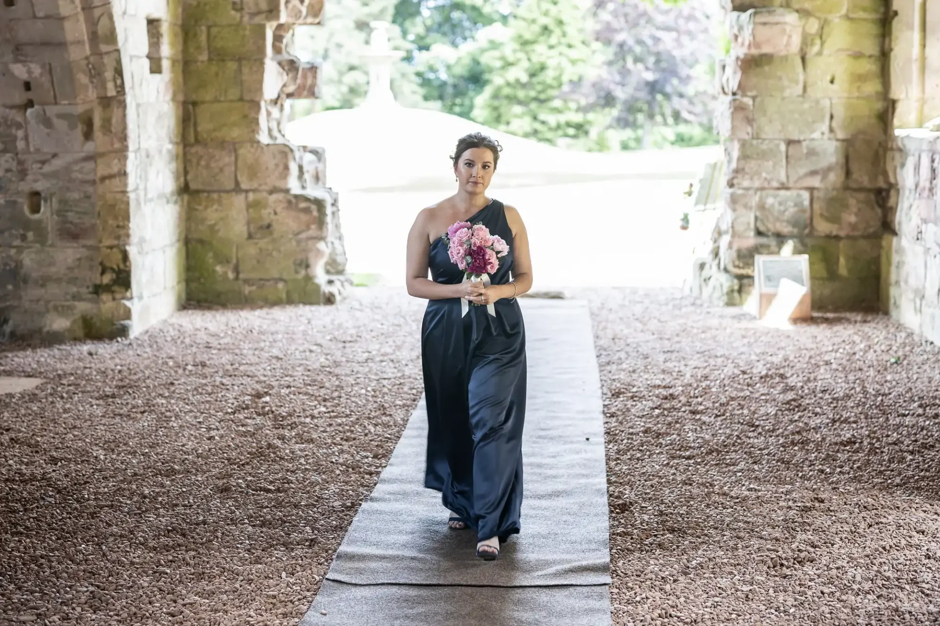 A woman in a navy dress walks down a stone aisle holding a bouquet, in a historic building with arches.