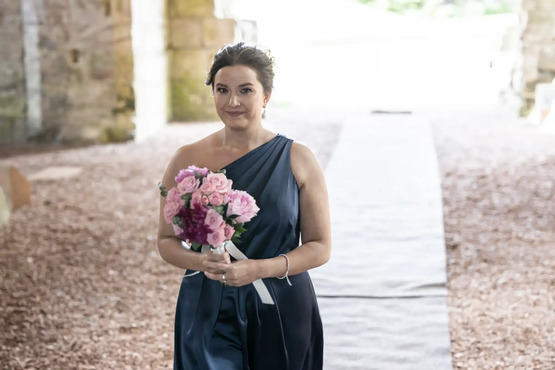 A woman in a navy blue dress holding a bouquet of pink flowers walks down a covered path with a serene expression.