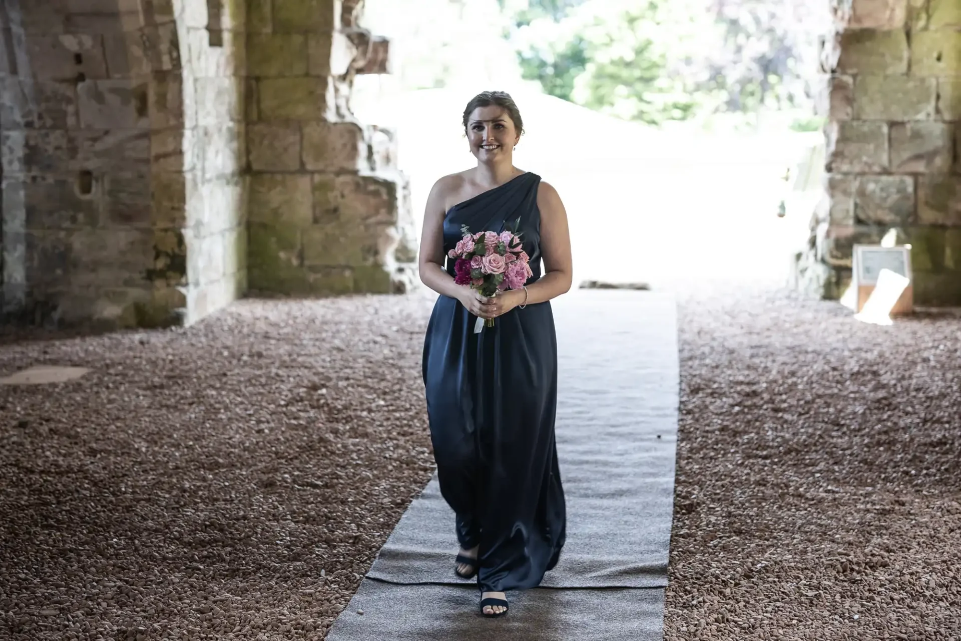 A woman in a navy blue gown holding a bouquet of pink flowers, smiling as she stands on a covered stone pathway.