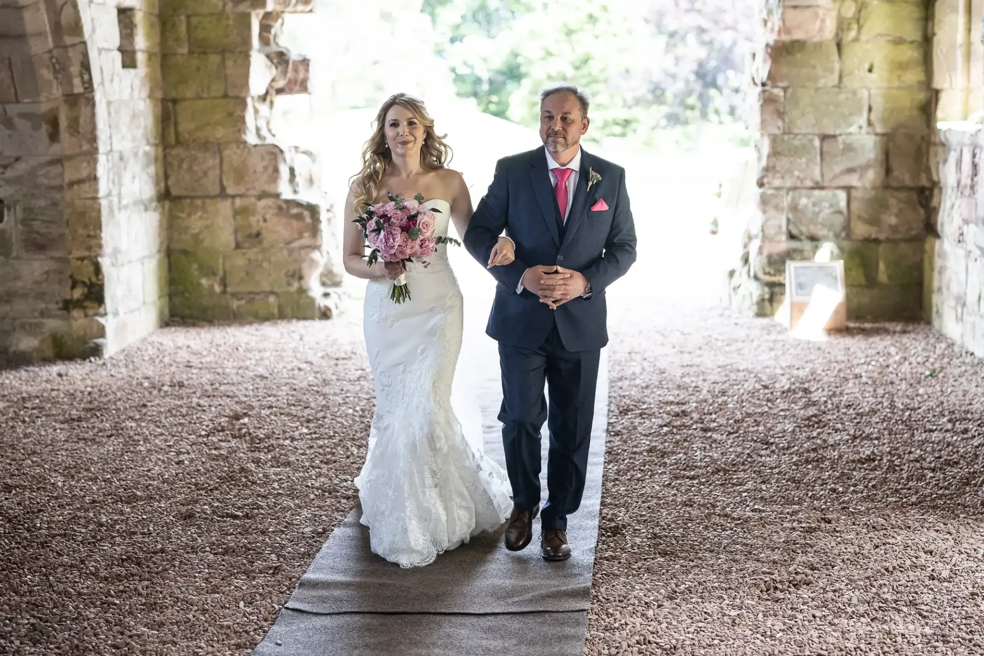 A bride in a white dress and a groom in a suit walk down the aisle together in a stone architecture setting, both smiling.