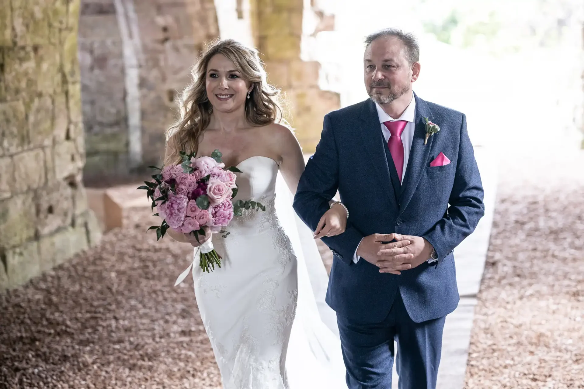 A bride in a white gown walks arm in arm with an older man in a suit, both smiling, under a stone archway, the bride holding a bouquet of pink flowers.