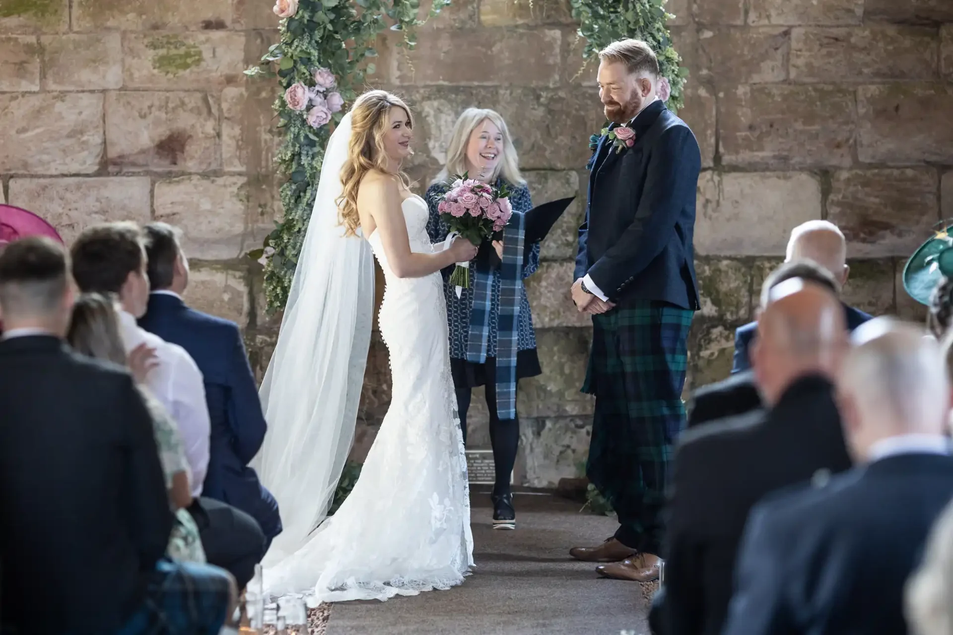 A bride and groom exchanging vows at their wedding ceremony, officiated by a woman, with guests in formal attire watching in a stone-walled venue decorated with flowers.