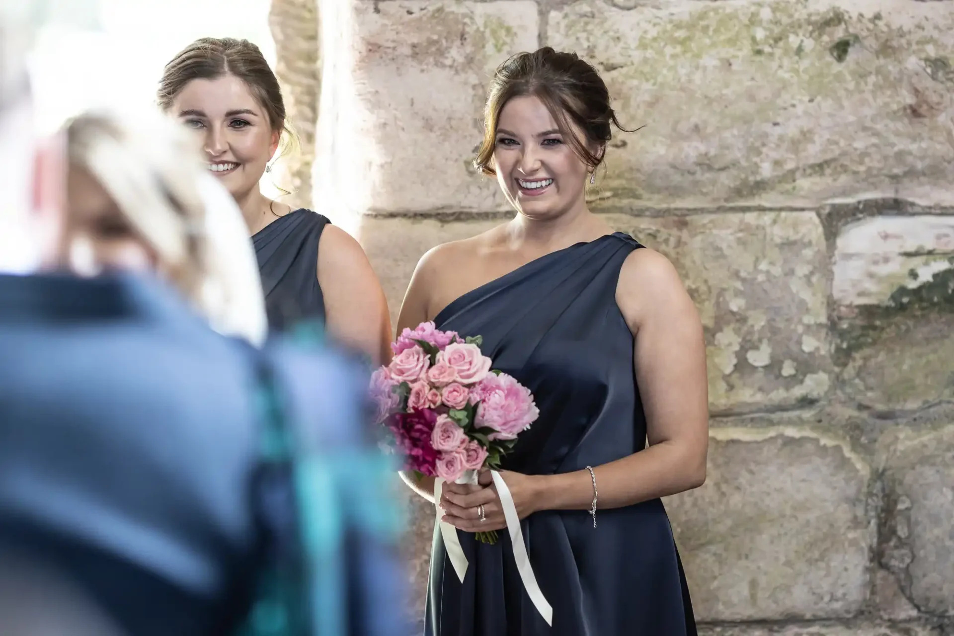 Two women in navy blue dresses smiling at a wedding, one holding a bouquet of pink roses.
