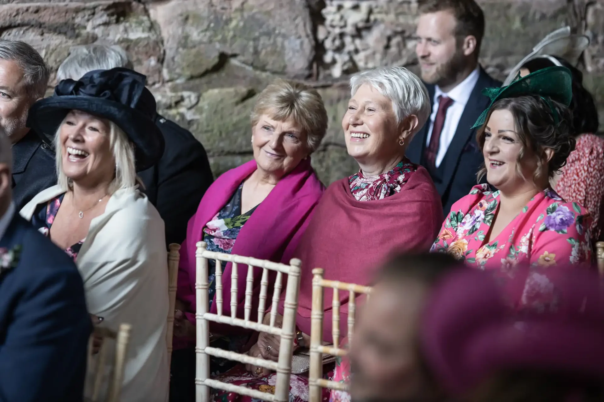 Group of elegantly dressed guests smiling and chatting at a wedding ceremony, held in a rustic stone venue.