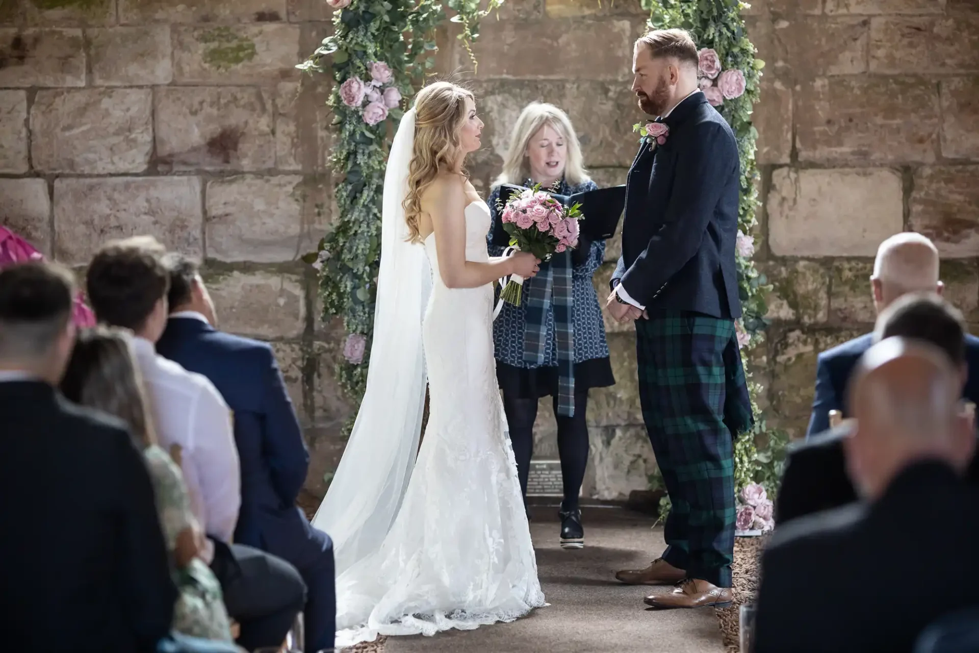 Bride in a white dress and groom in a kilt exchanging vows at a wedding ceremony, surrounded by guests and floral decorations.