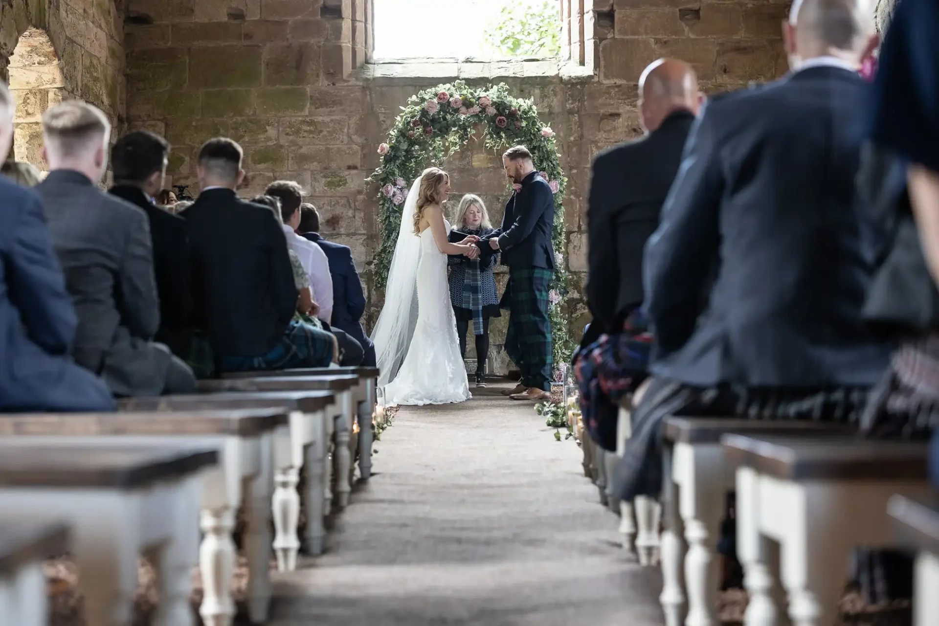 A bride and groom holding hands during their wedding ceremony in a rustic church setting, surrounded by guests.