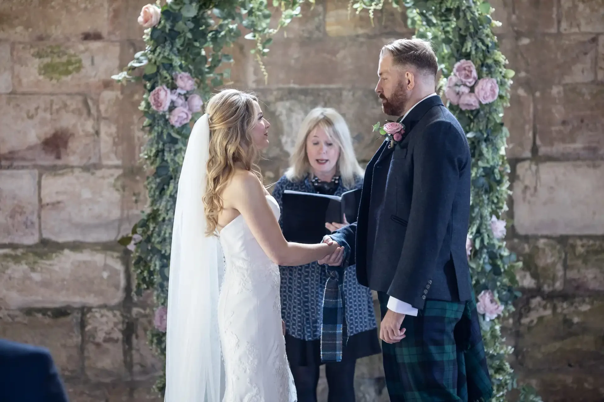 A bride and groom hold hands during their wedding ceremony, officiated by a woman, in a venue decorated with roses and ivy. the groom wears a kilt.