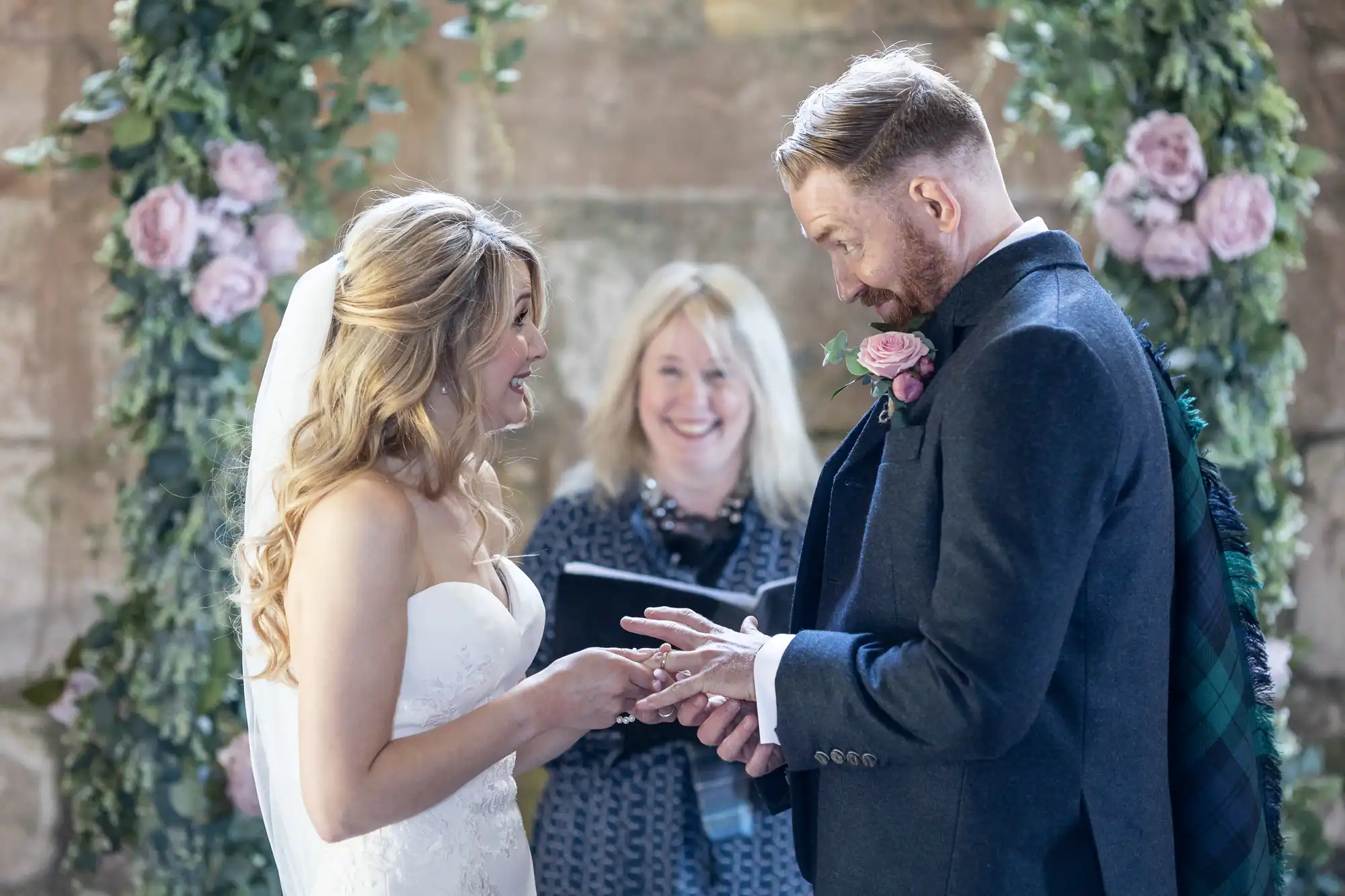 Bride and groom exchanging rings during a wedding ceremony, with an officiant smiling in the background, in a venue decorated with flowers.