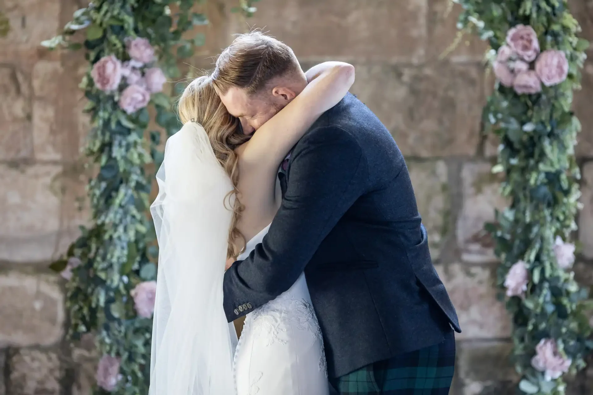 A bride and groom embracing at their wedding, the groom wearing a kilt, surrounded by floral decorations in a stone building.