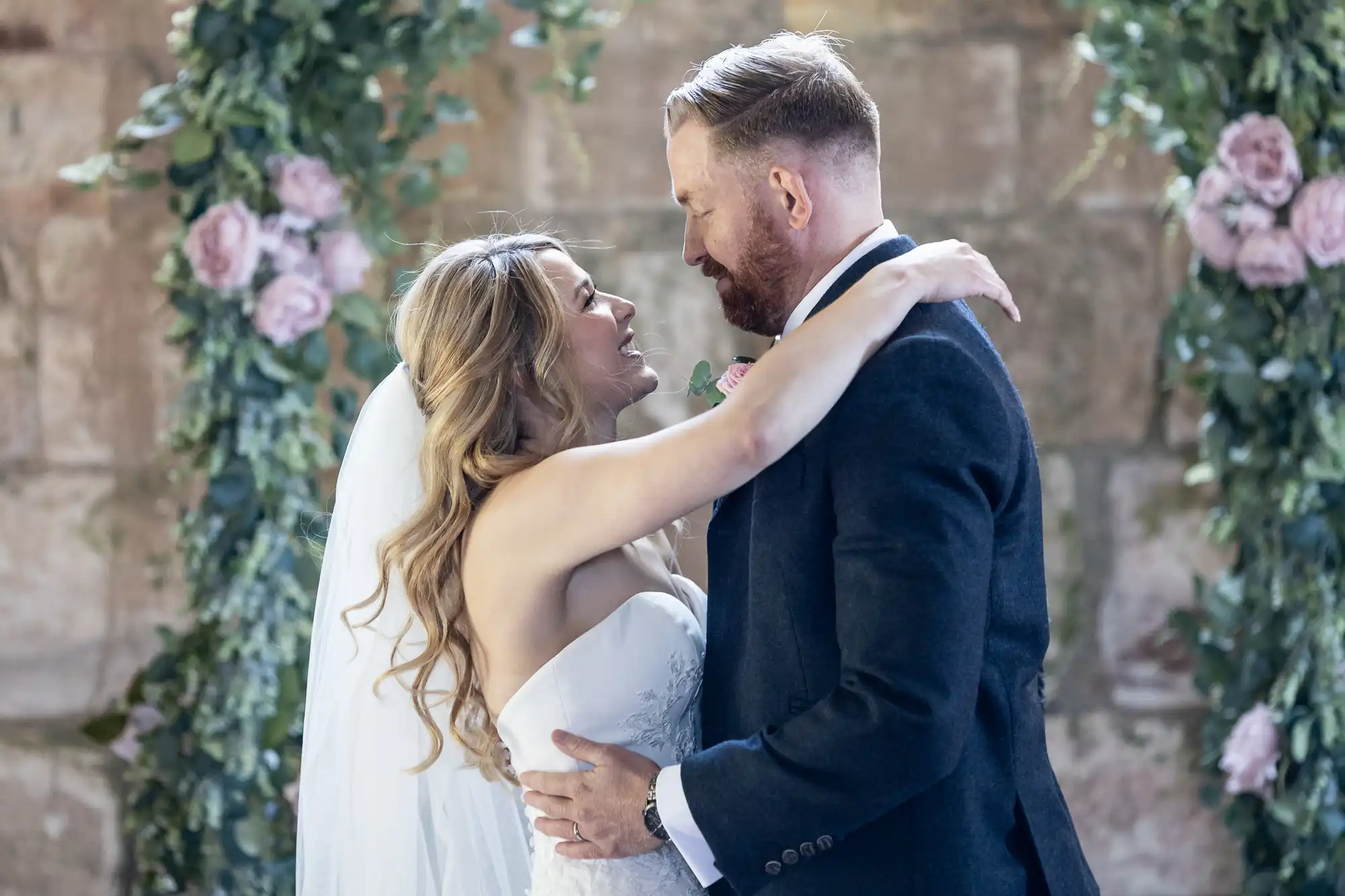 A bride and groom embrace and gaze into each other's eyes, surrounded by pink roses in an old stone building.